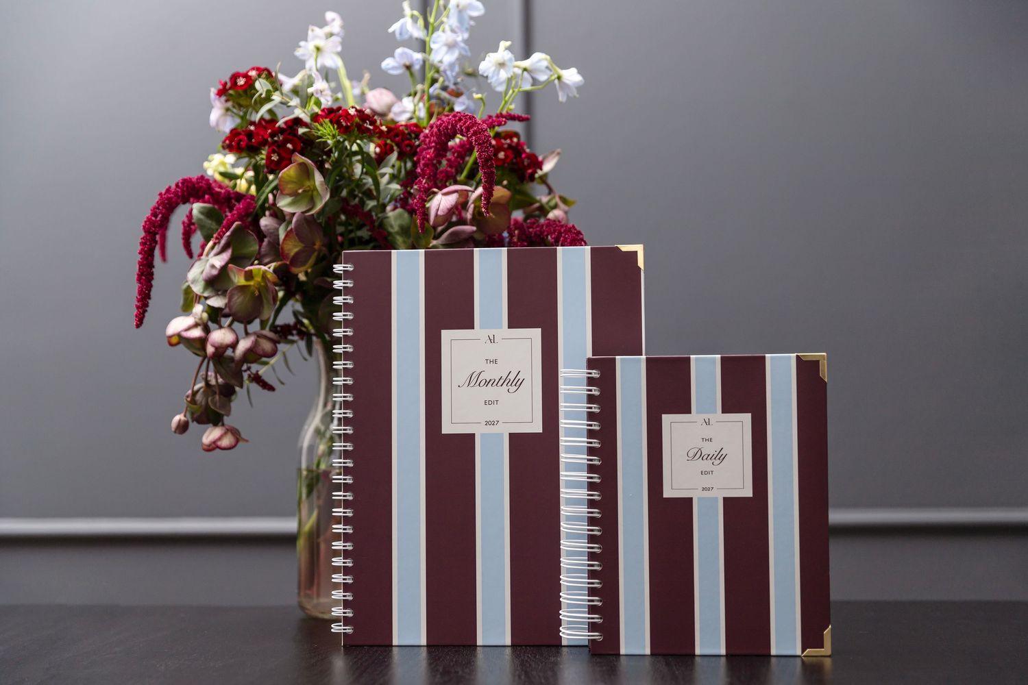 Two striped burgundy and white spiral notebooks beside a floral arrangement on a dark surface.