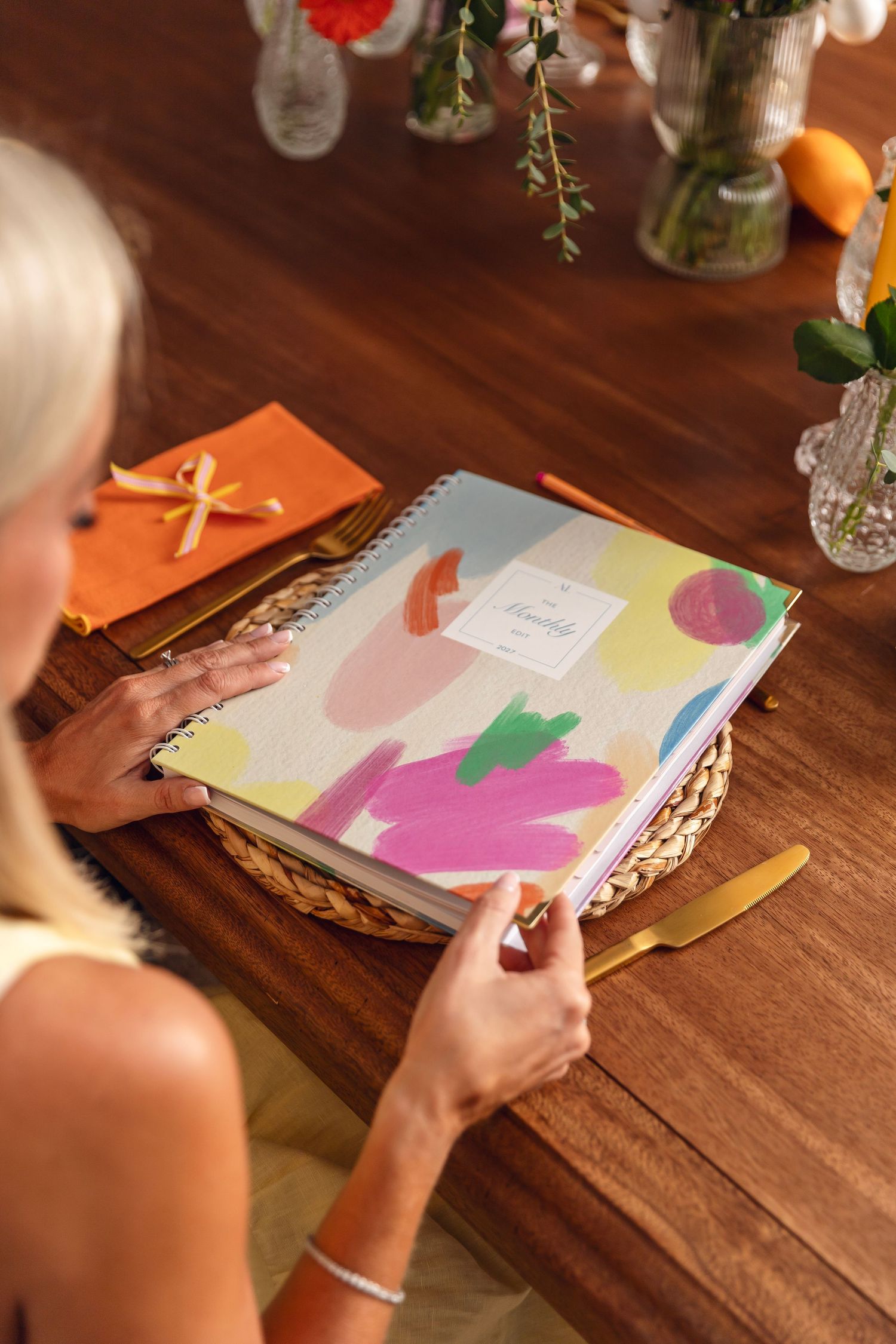 Blonde woman holding a colorful spiral-bound planner at a wooden table with gold cutlery and floral decor.