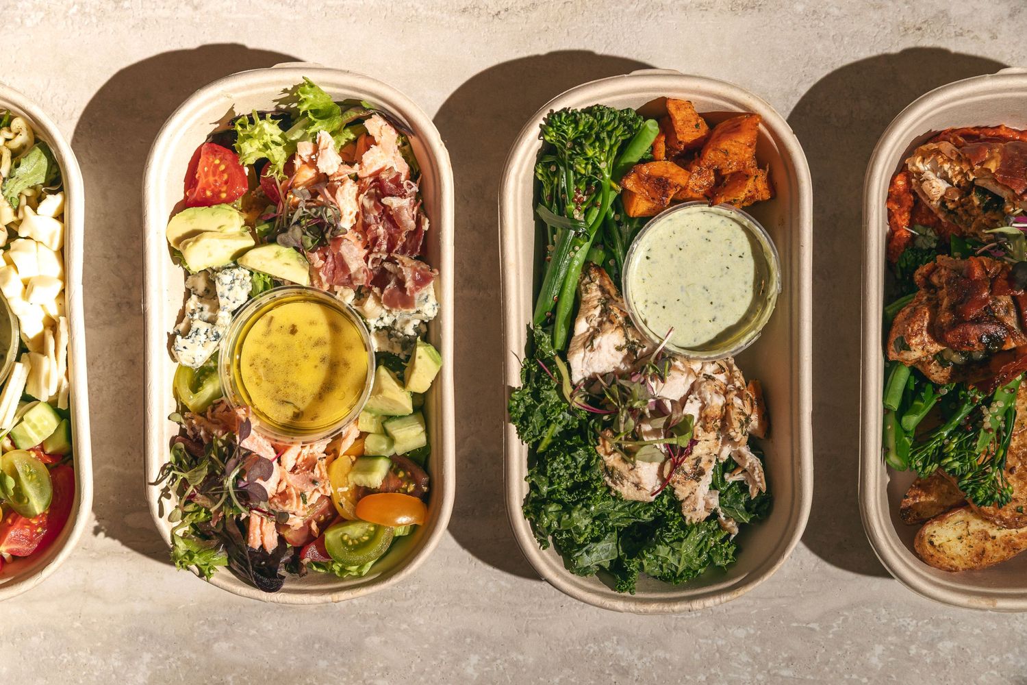 Four oval meal prep containers with fresh salads, roasted vegetables, grilled chicken, and dipping sauces on a light surface.
