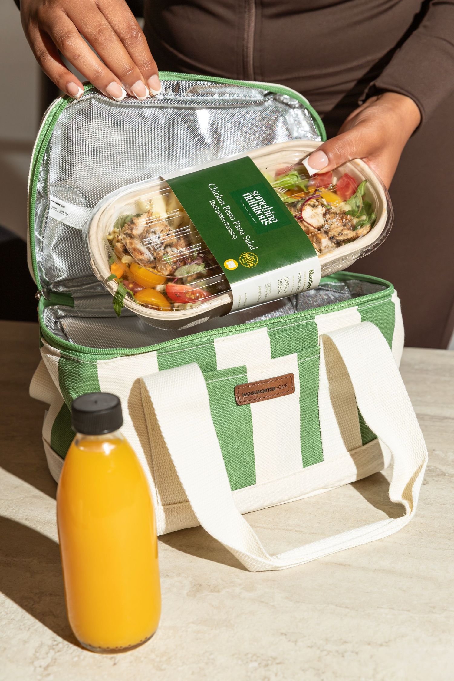 Person placing a healthy meal prep container into a green and white insulated lunch bag with an orange juice bottle.
