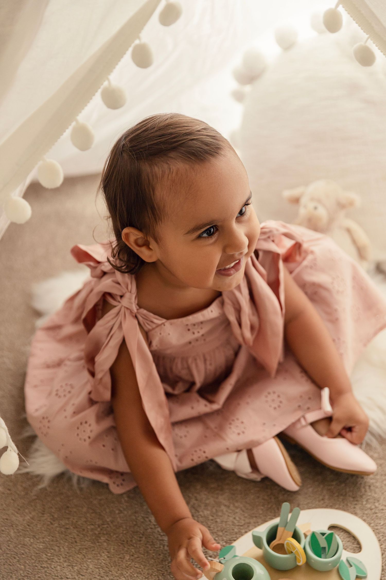 Baby in pink lacy dress lying on carpet reaching for colorful wooden toys during playtime.