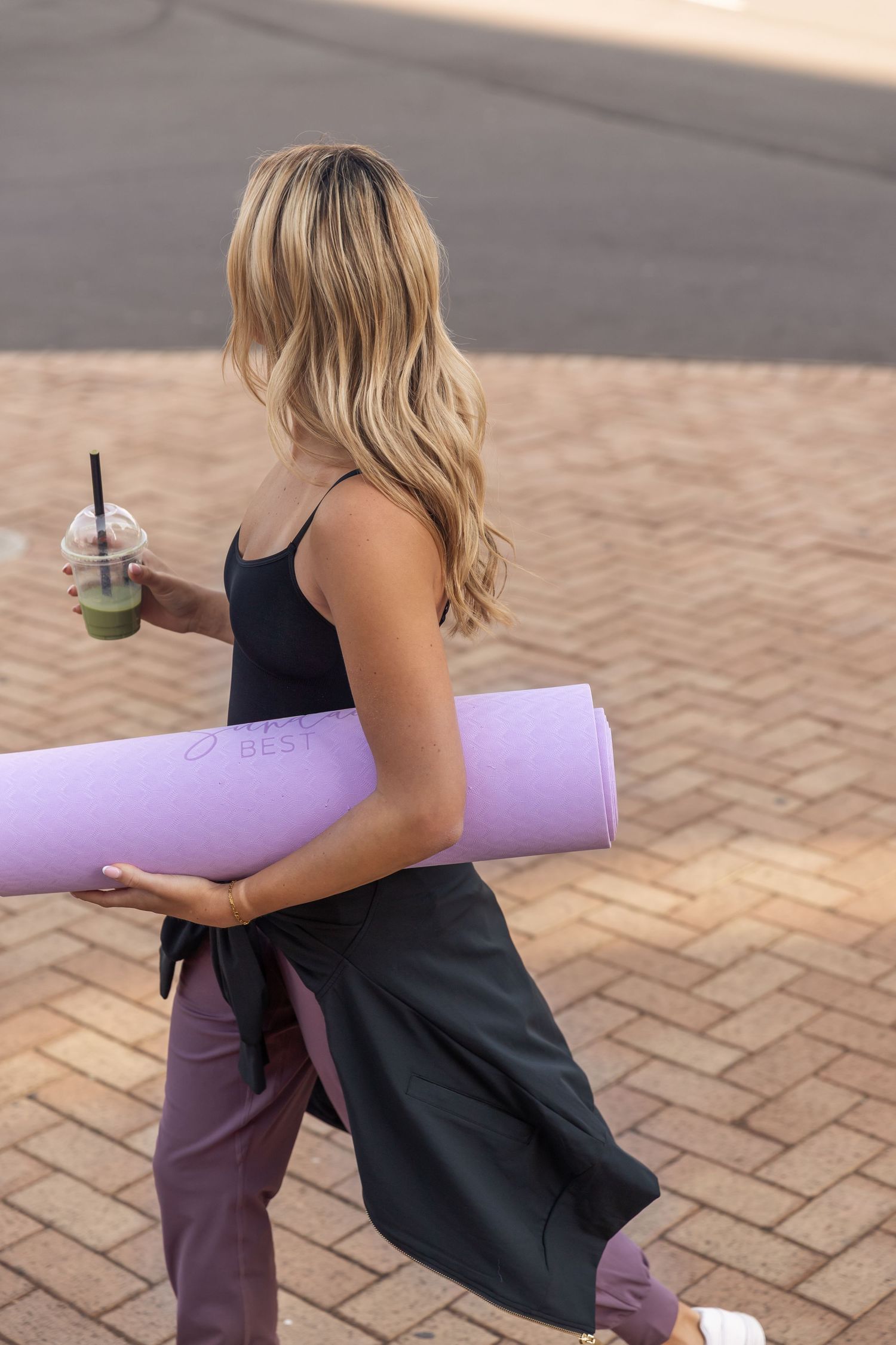 A person in black athletic wear rolls up a purple yoga mat while holding a green smoothie on outdoor brick pavement.