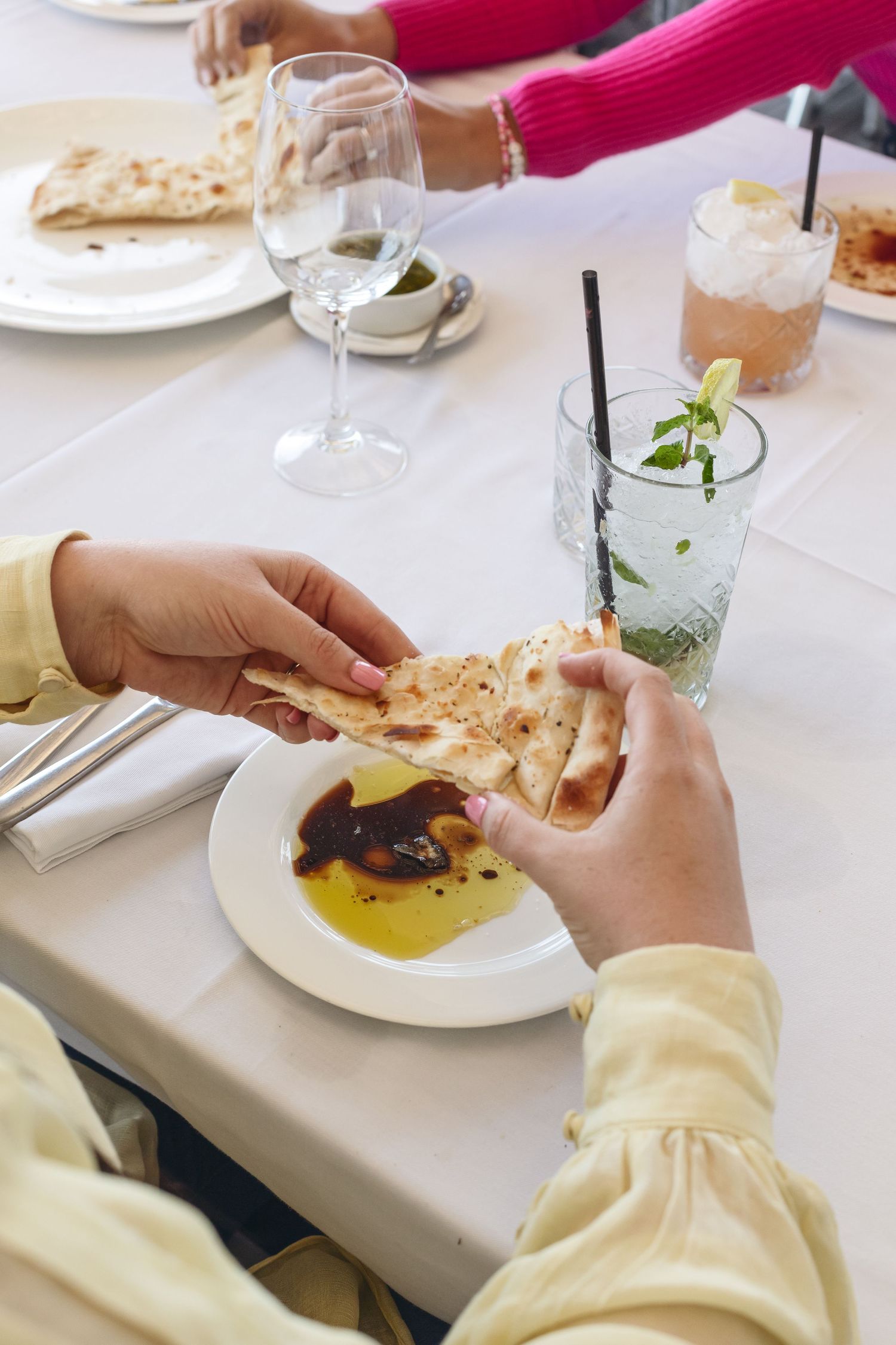 Hands tearing pita bread over a plate of olive oil dip at an elegant restaurant table setting with cocktails.