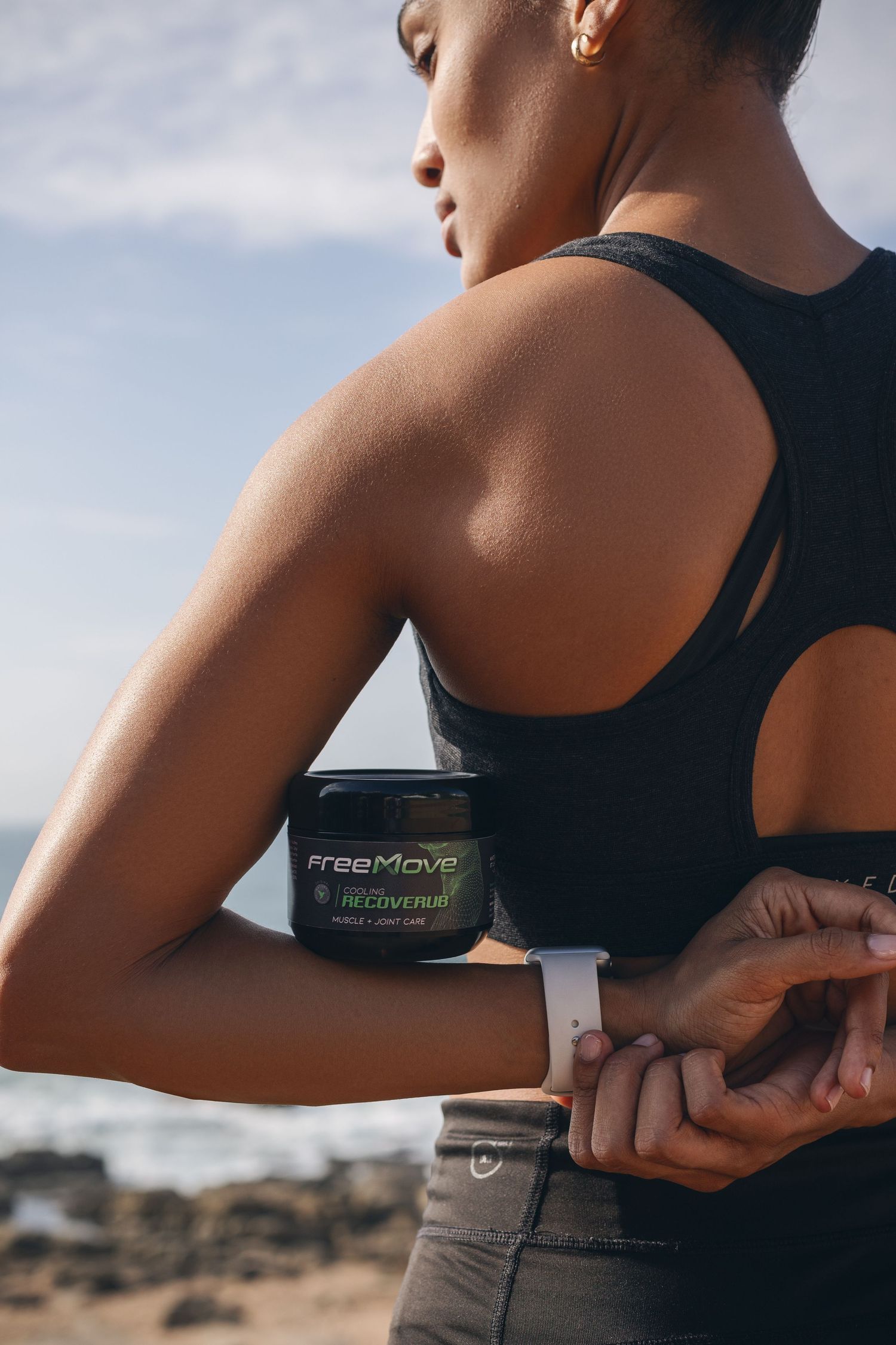 Athletic person in black sports bra applies FreeMove joint cream outdoors against a rocky landscape backdrop.
