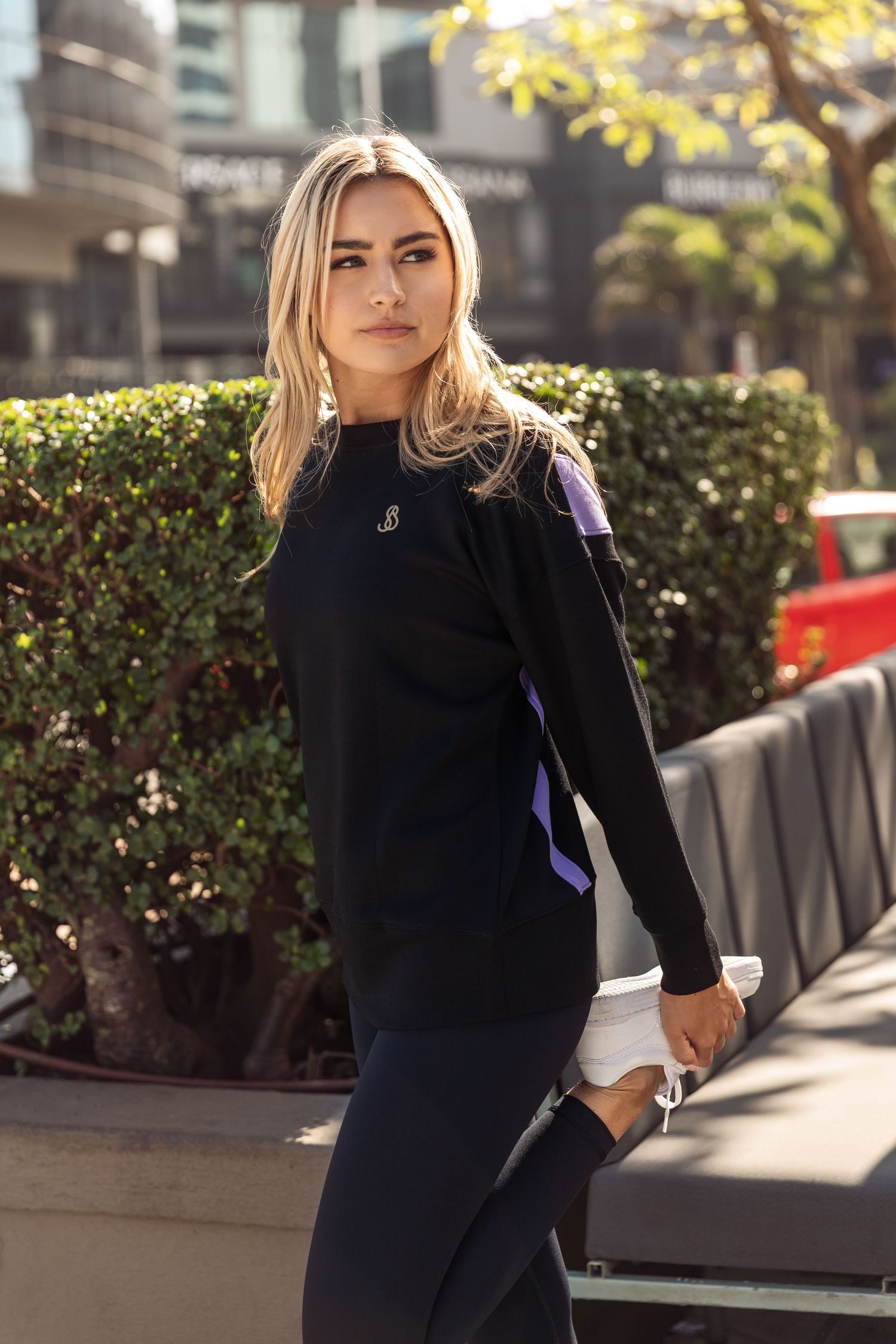 Person in a black sweater and dark pants poses outdoors on a sunny day with urban greenery in the background.