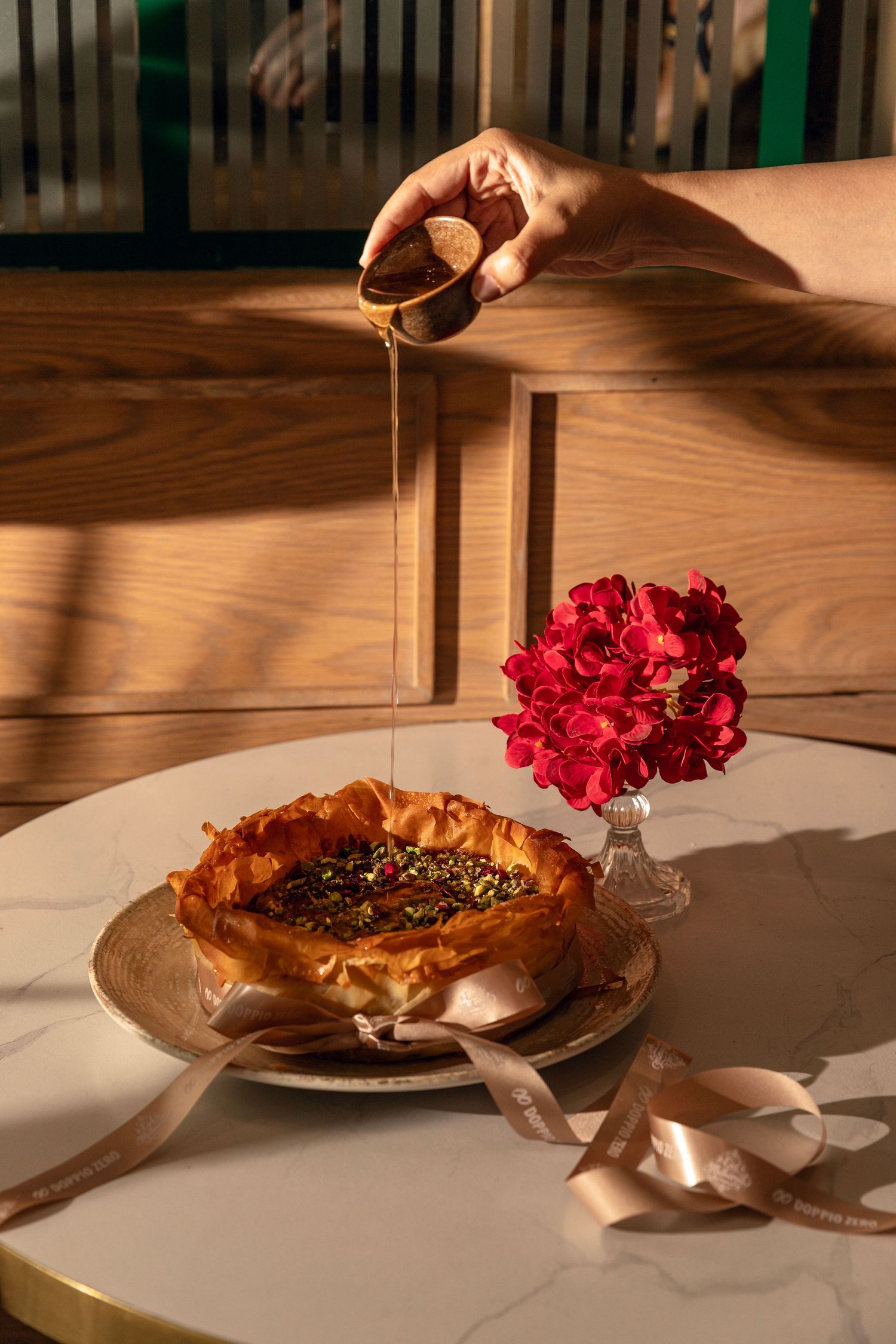 A hand drizzles honey over a rustic pastry on a plate, with a red carnation flower arrangement and pink ribbon nearby.