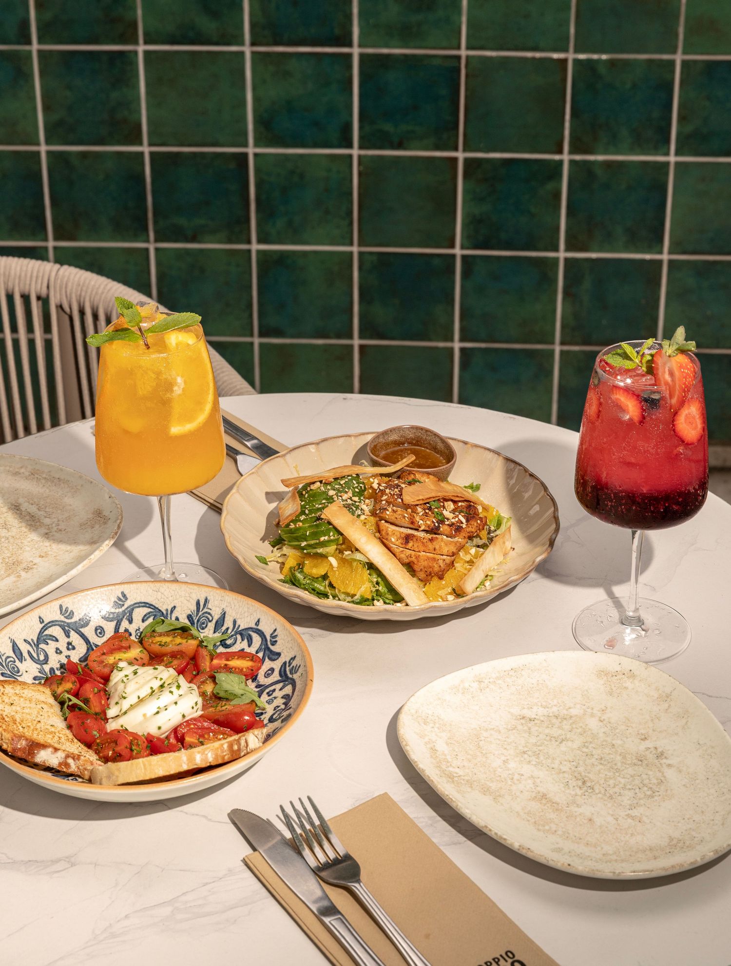 A restaurant table setting with colorful cocktails, flatbread, and plates against green tiled wall in natural lighting.