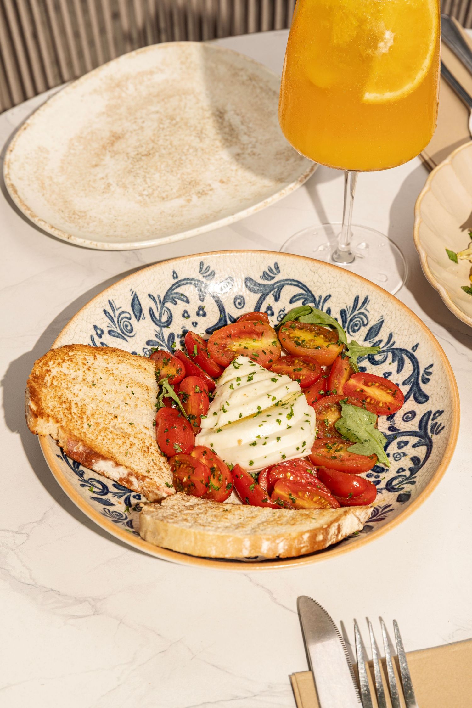 A plate of burrata salad with tomatoes and toasted bread served alongside a glass of orange juice on a white table.