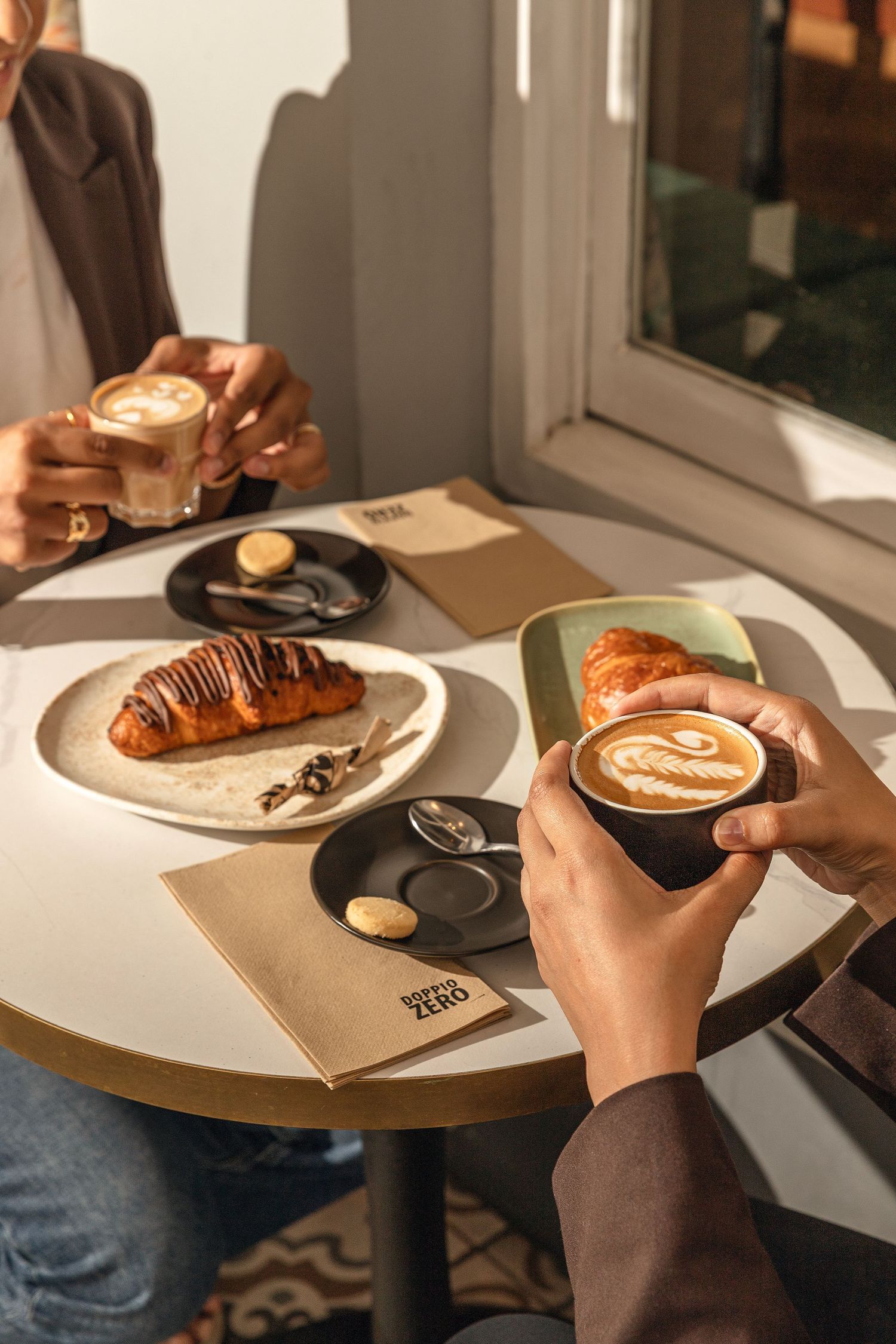 Hands holding coffee cups and pastries on a round cafe table with brown paper placemats in warm lighting.