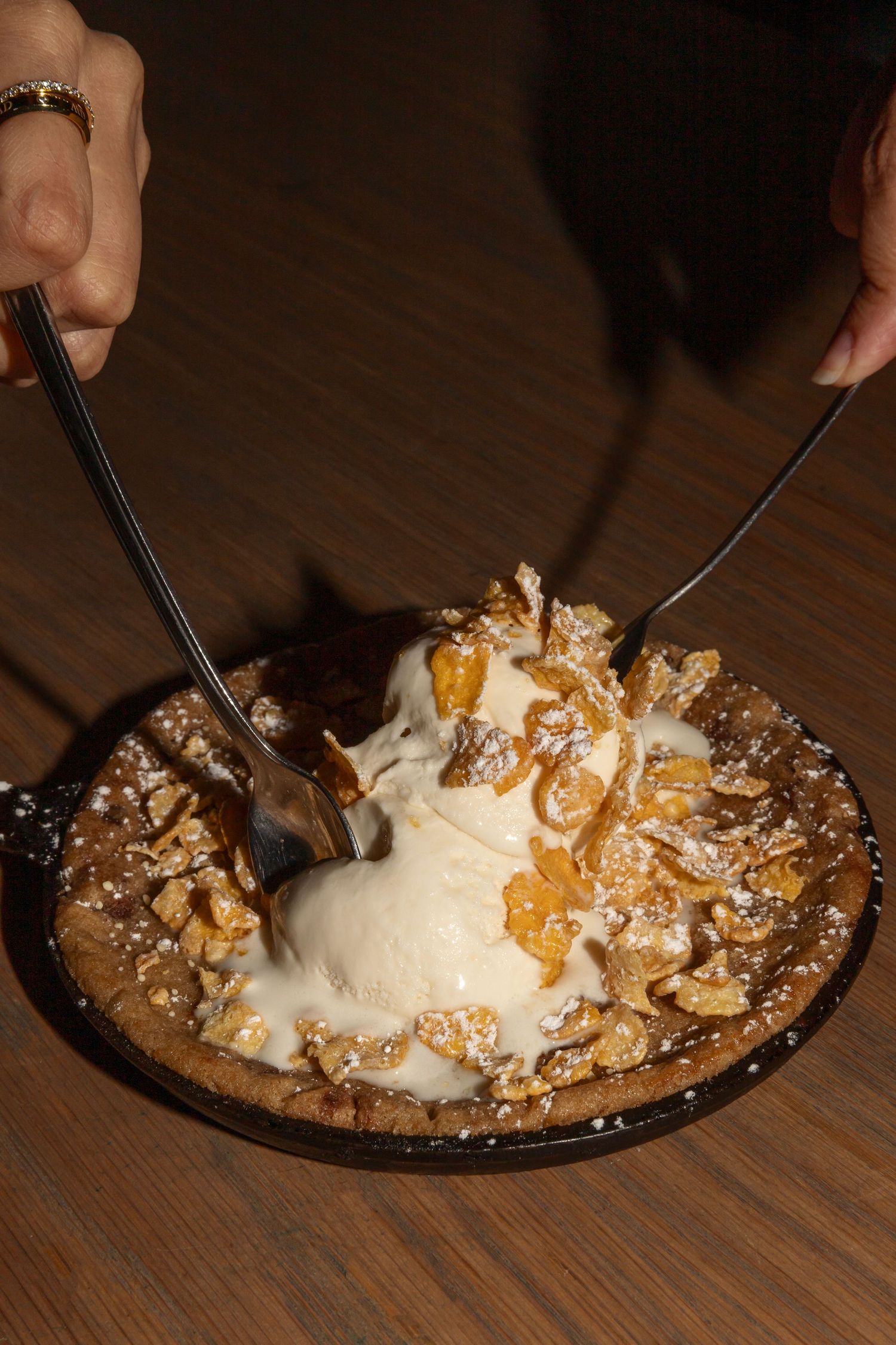 A skillet dessert with vanilla ice cream, candied nuts, and powdered sugar being served by two hands.