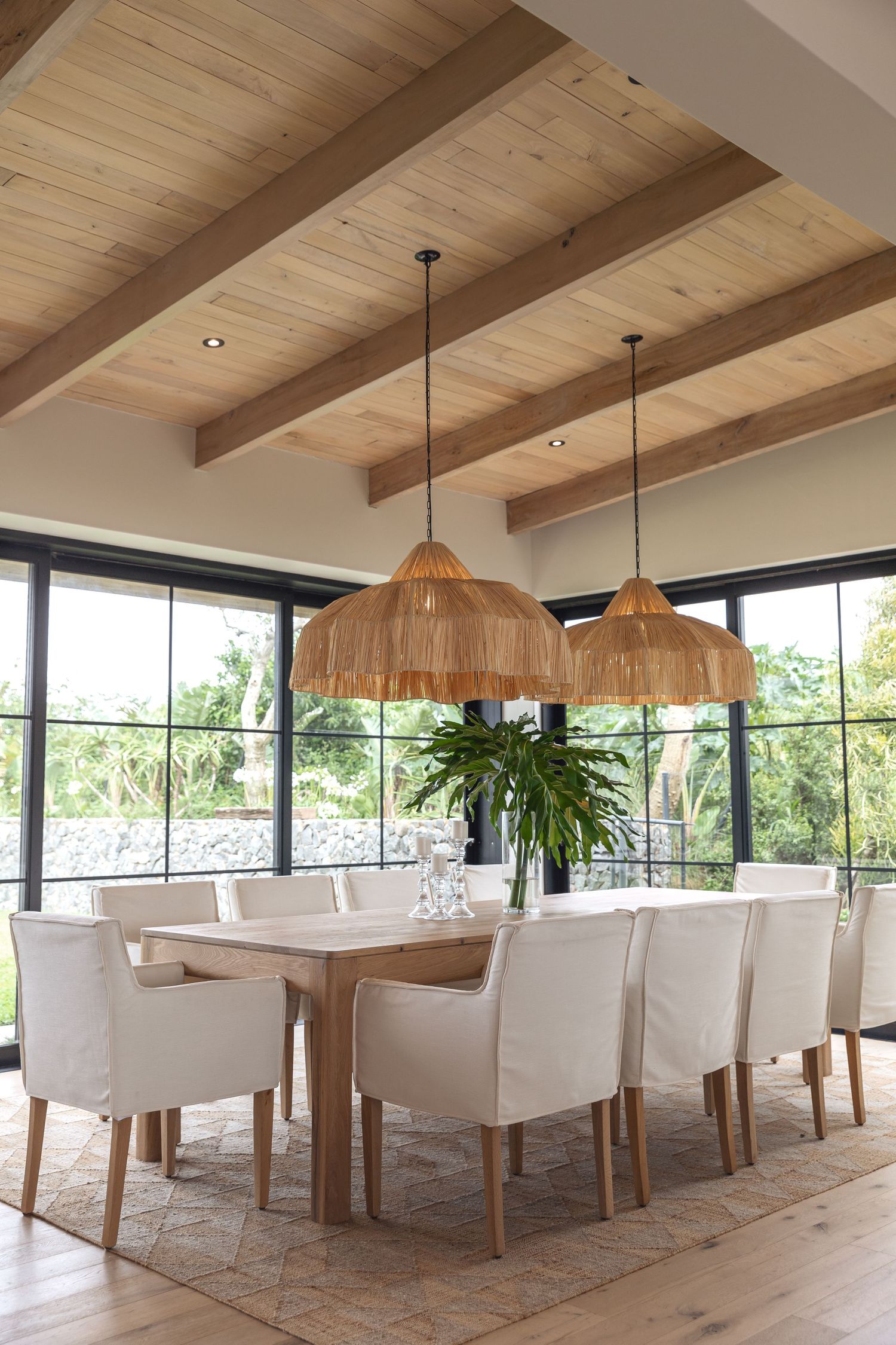 Modern dining room with wooden beamed ceiling, woven pendant lights, and white upholstered chairs around a wooden table.