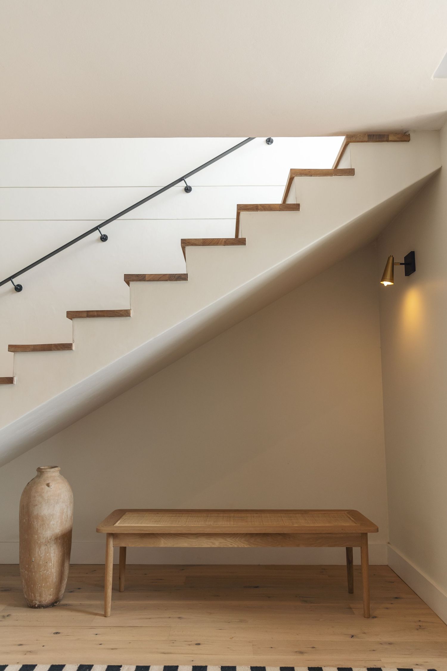 Modern interior staircase with wooden treads, metal handrail, and simple wooden bench beneath against a neutral wall.