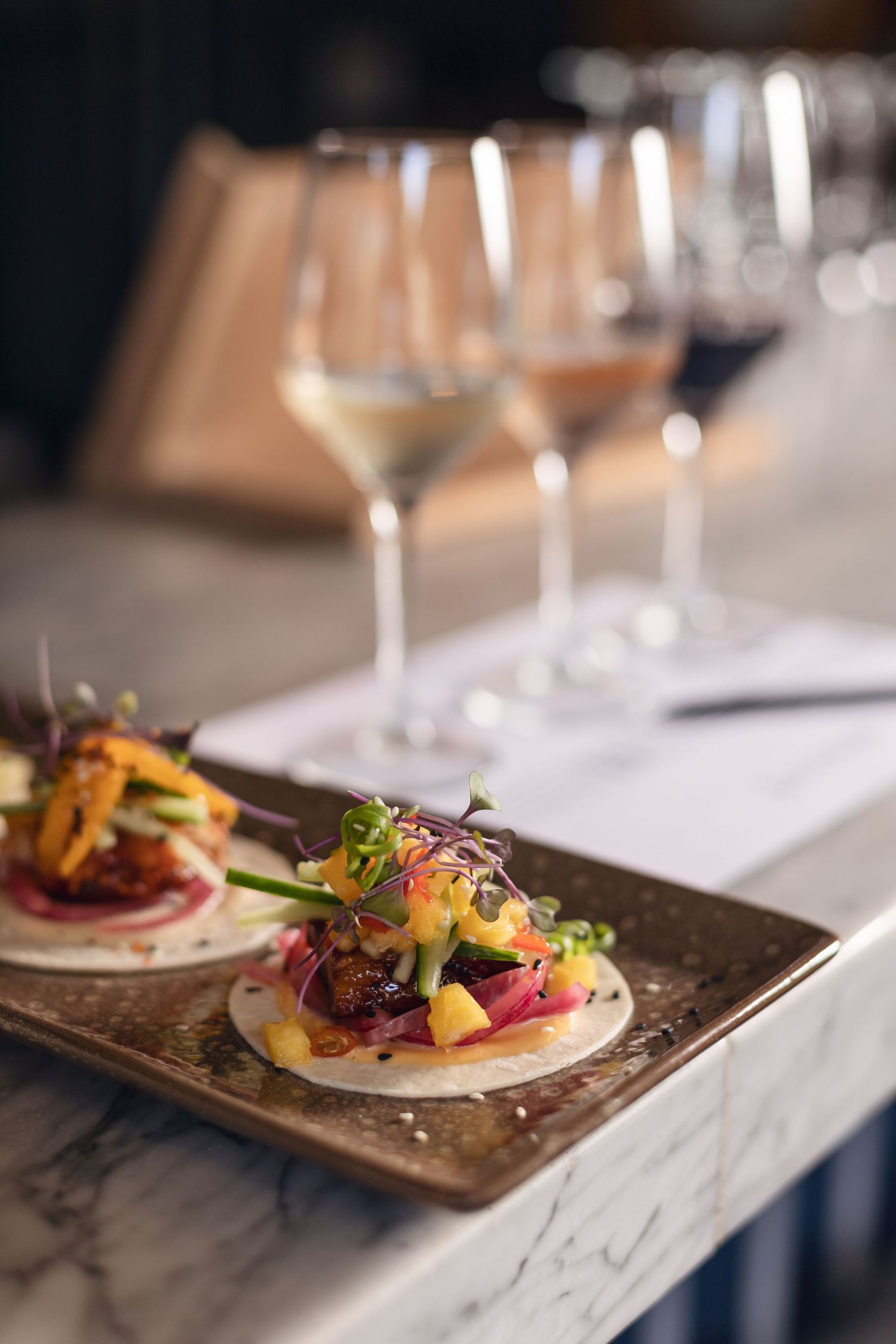 Elegant taco appetizers with colorful toppings served on a stone plate with wine glasses on marble counter.