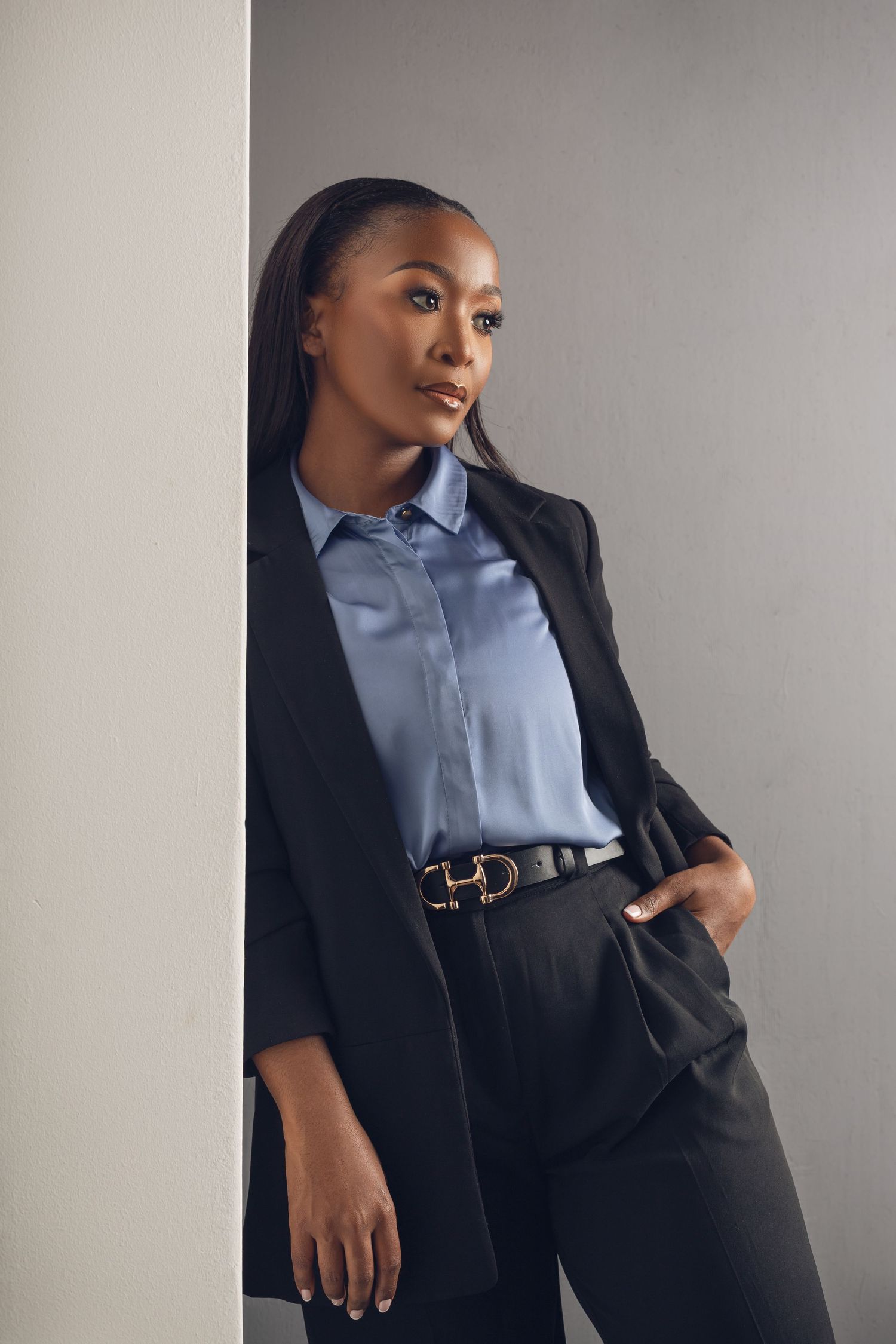 Professional business attire featuring a black blazer, blue button-down shirt, and sleek leather belt against a gray backdrop.