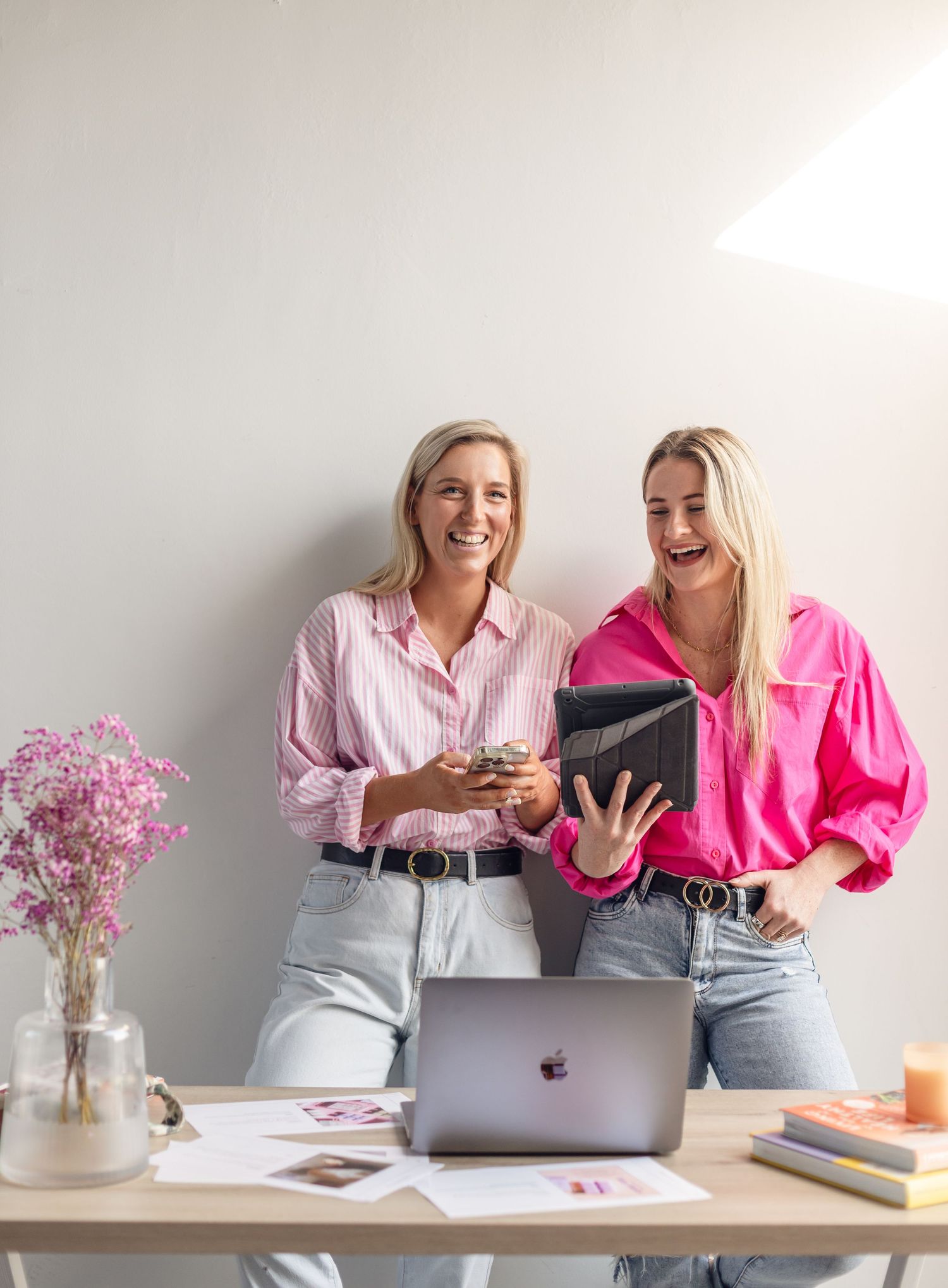 Two colleagues share a laugh while looking at digital devices and working at a desk decorated with pink flowers.