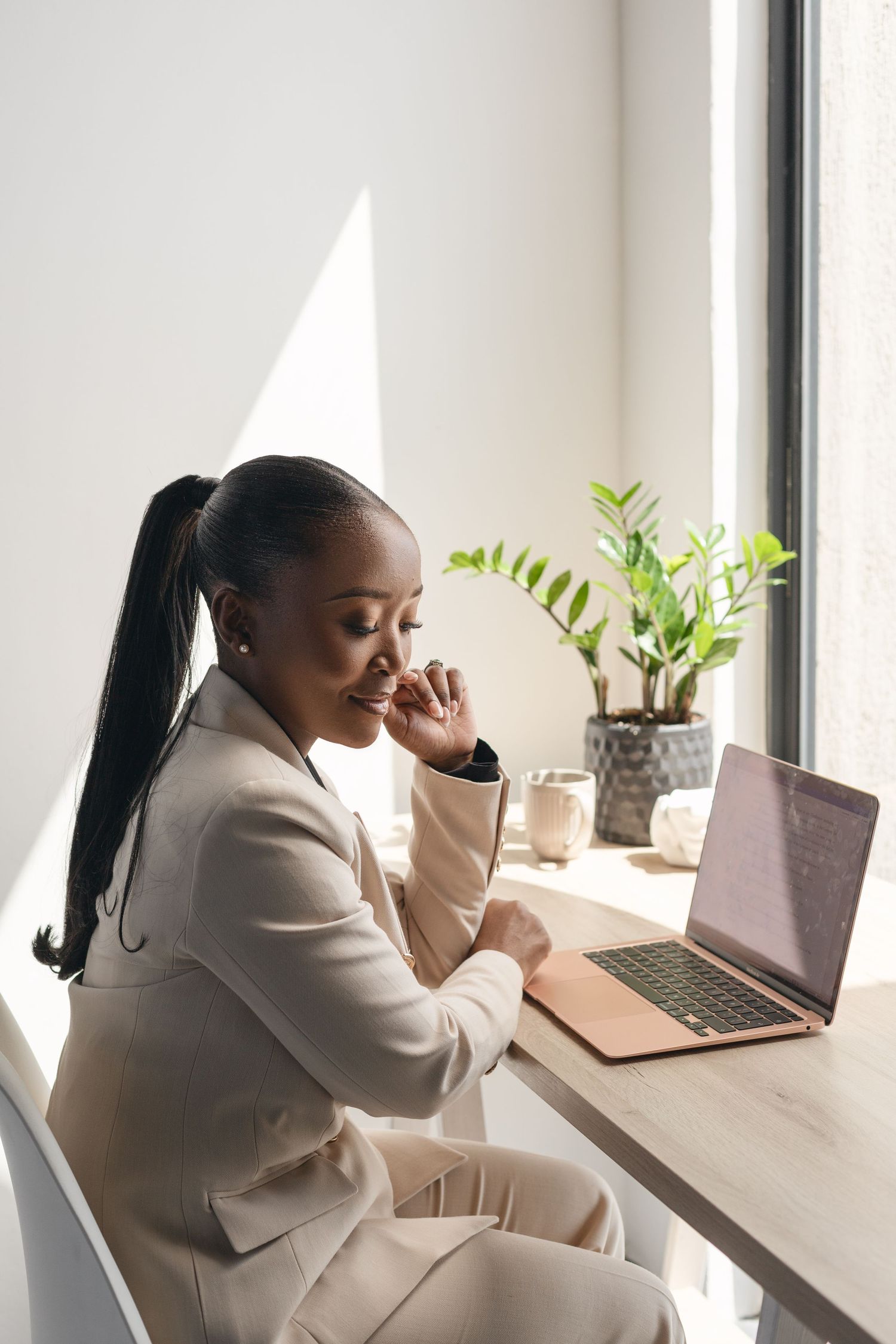 Professional in a beige suit sitting at desk with laptop and potted plant by window in bright modern office setting.