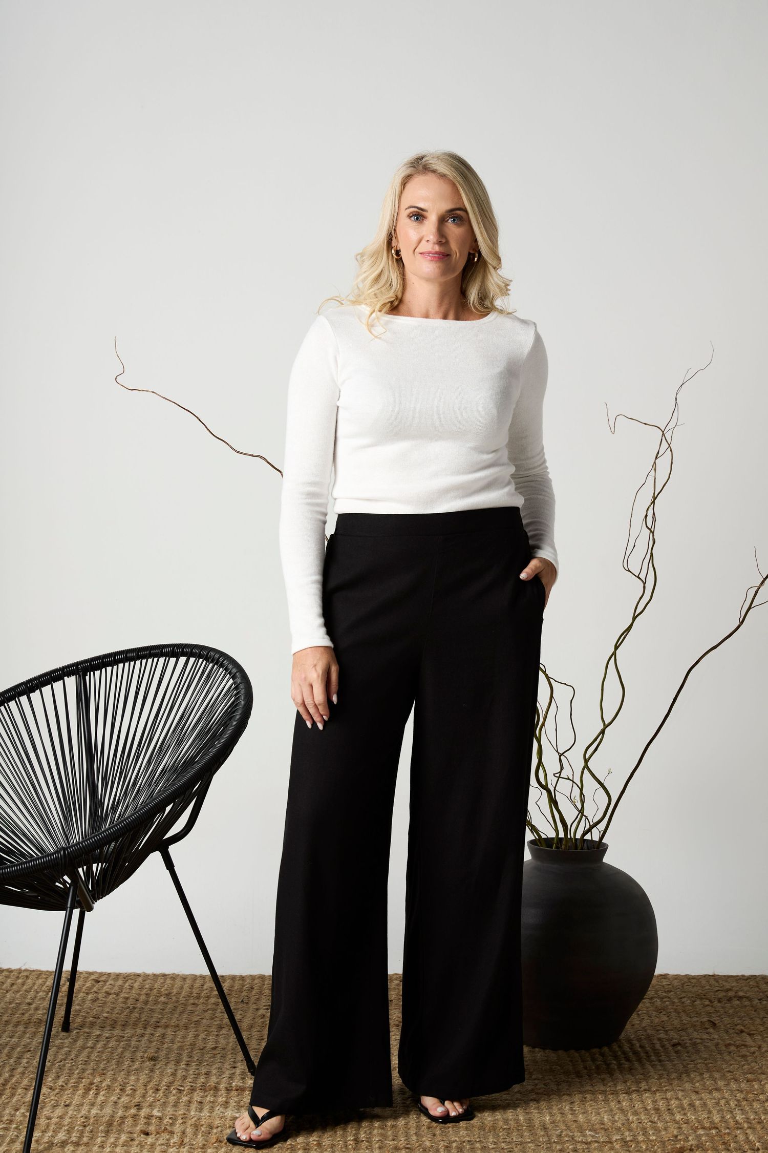 Woman in white long-sleeve top and black wide-leg trousers standing in a minimalist studio setting.