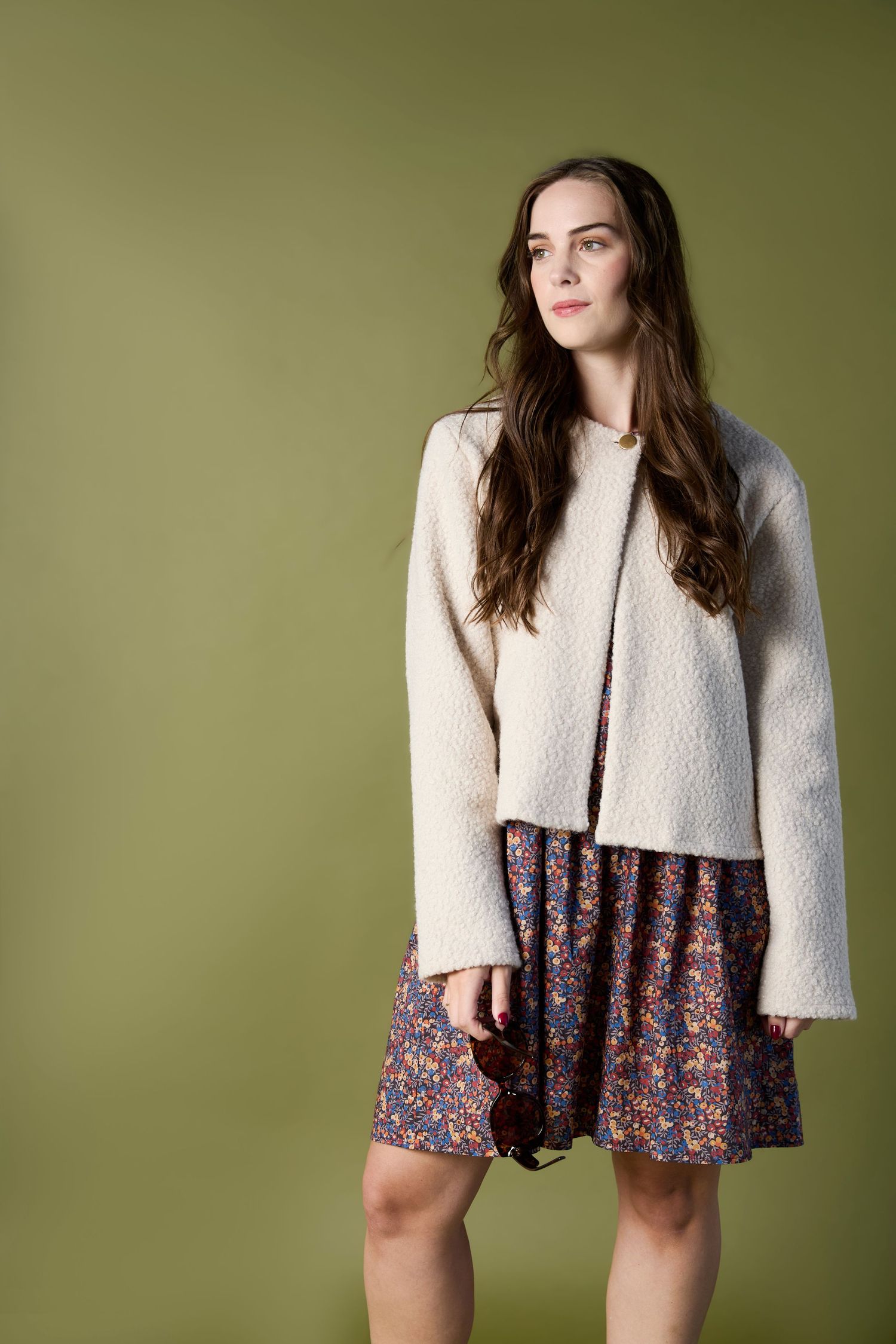 Woman in cream textured jacket and floral mini dress holding sunglasses against olive green background.