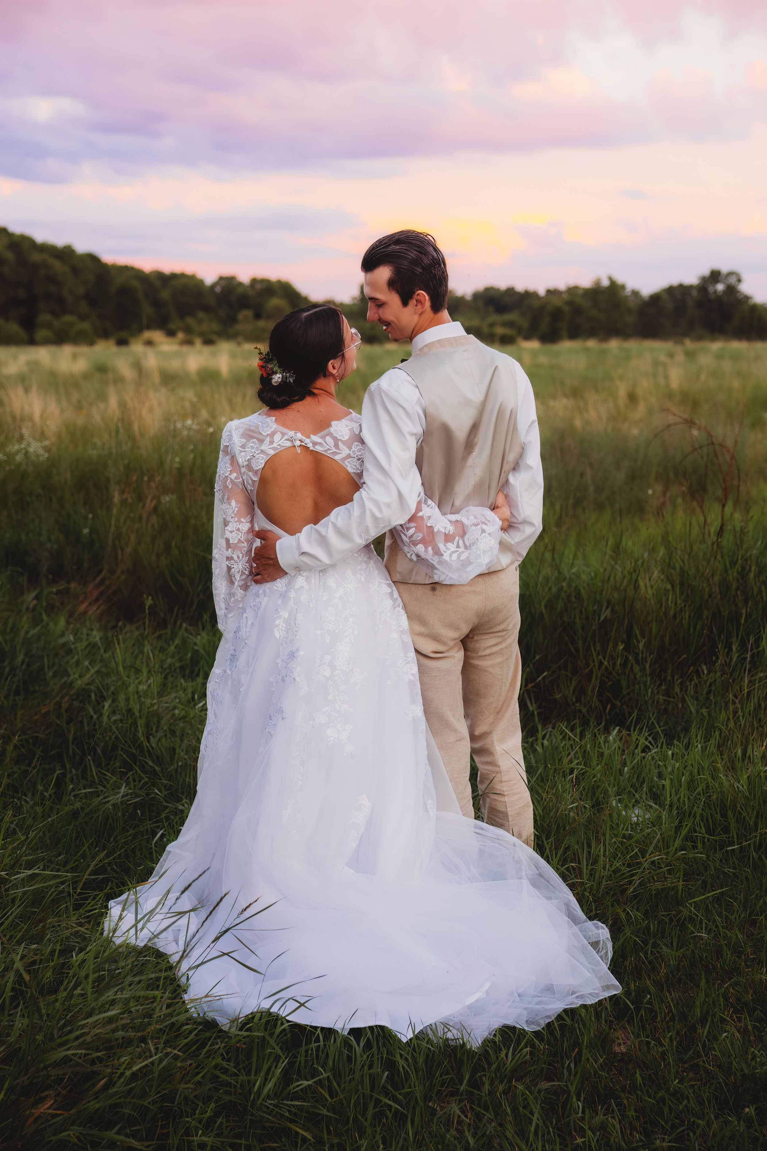 A romantic couple in wedding attire embraces while standing in a golden meadow at sunset.