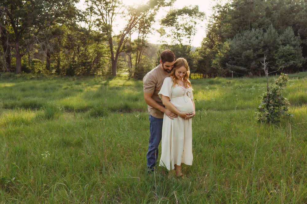 Couple embracing in a sunlit green meadow, woman in white maternity dress, man in jeans, golden hour outdoor pregnancy photo.