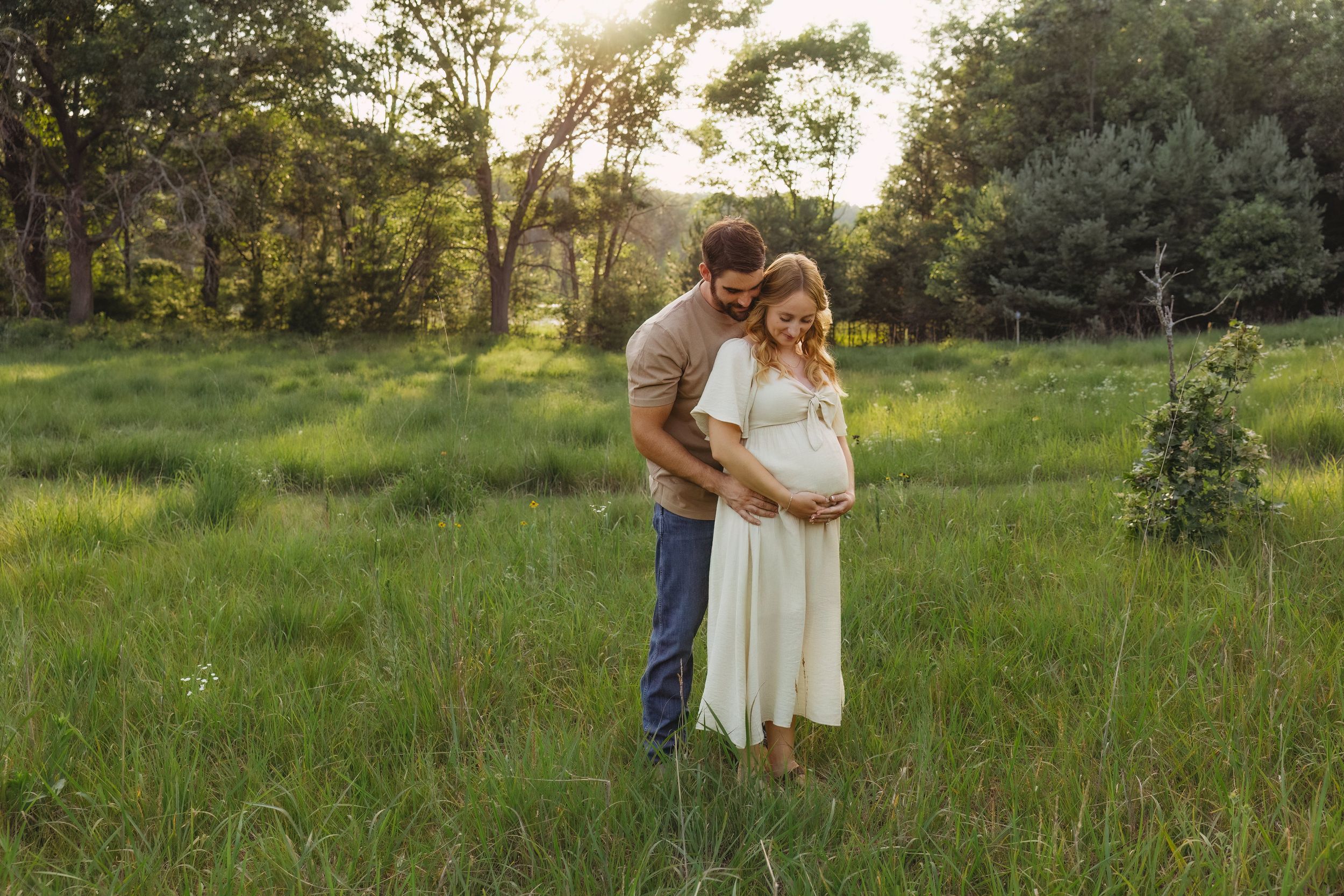 Couple embracing in a sunlit green meadow, woman in white maternity dress, man in jeans, golden hour outdoor pregnancy photo.