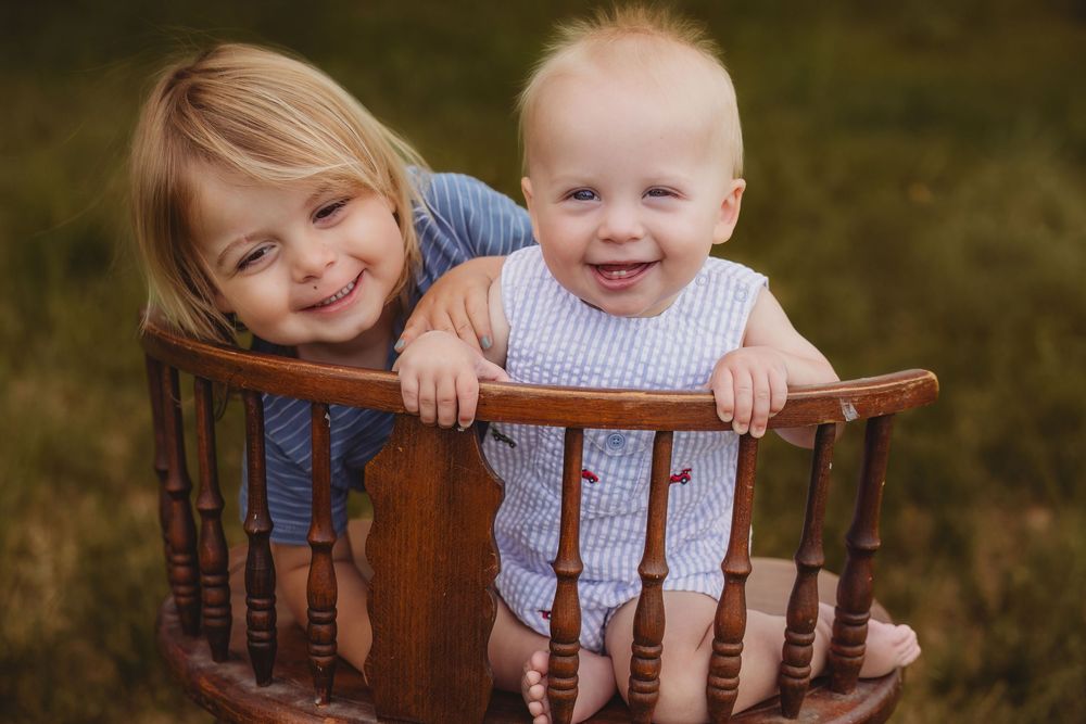 Two smiling young siblings, a toddler girl and baby boy, pose together in a wooden chair outdoors.