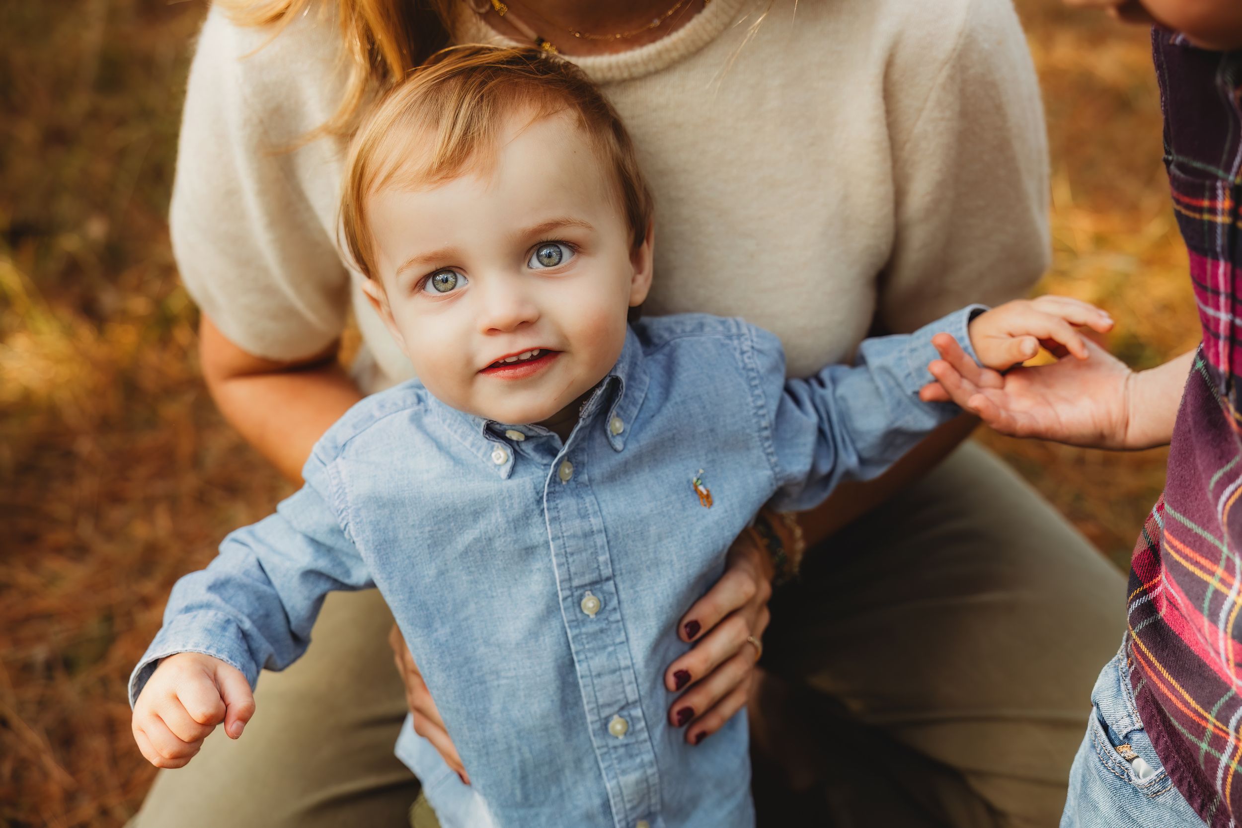 A child in a denim shirt smiles while being held outdoors during an autumn photo session.