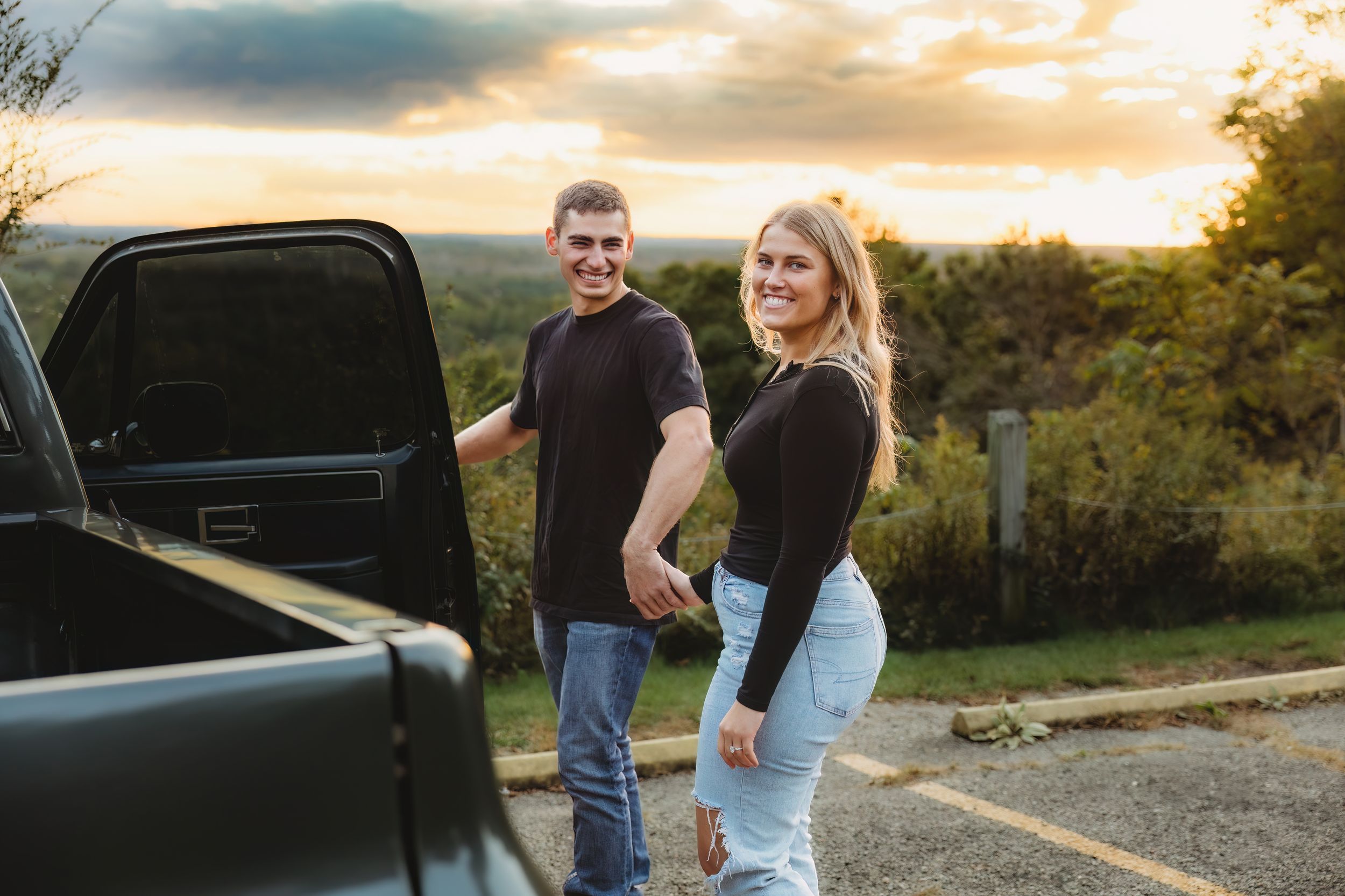 Two people stand next to a black pickup truck at sunset on a hilltop overlooking scenic countryside views.