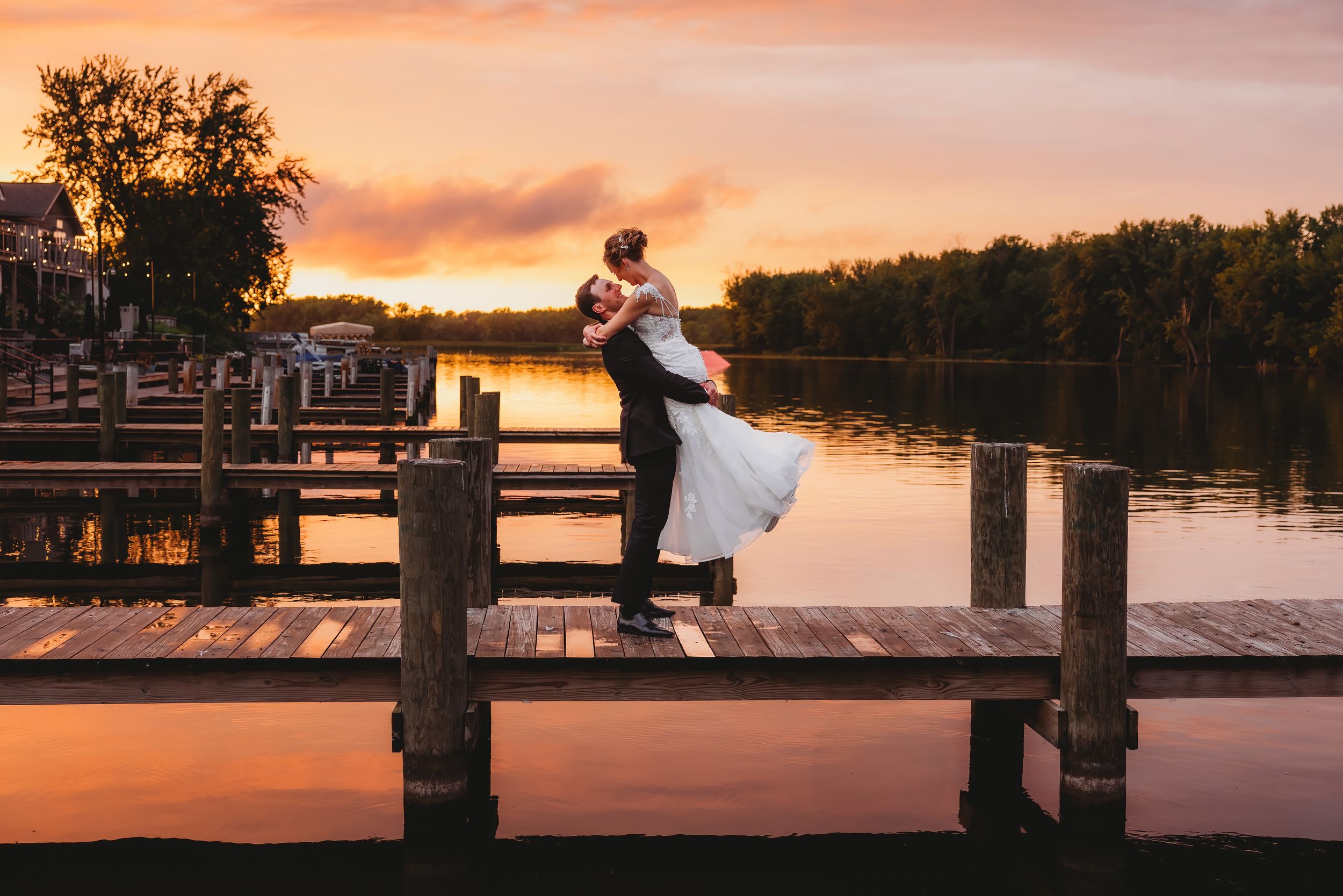 Romantic sunset wedding photo of couple embracing on wooden dock over lake.