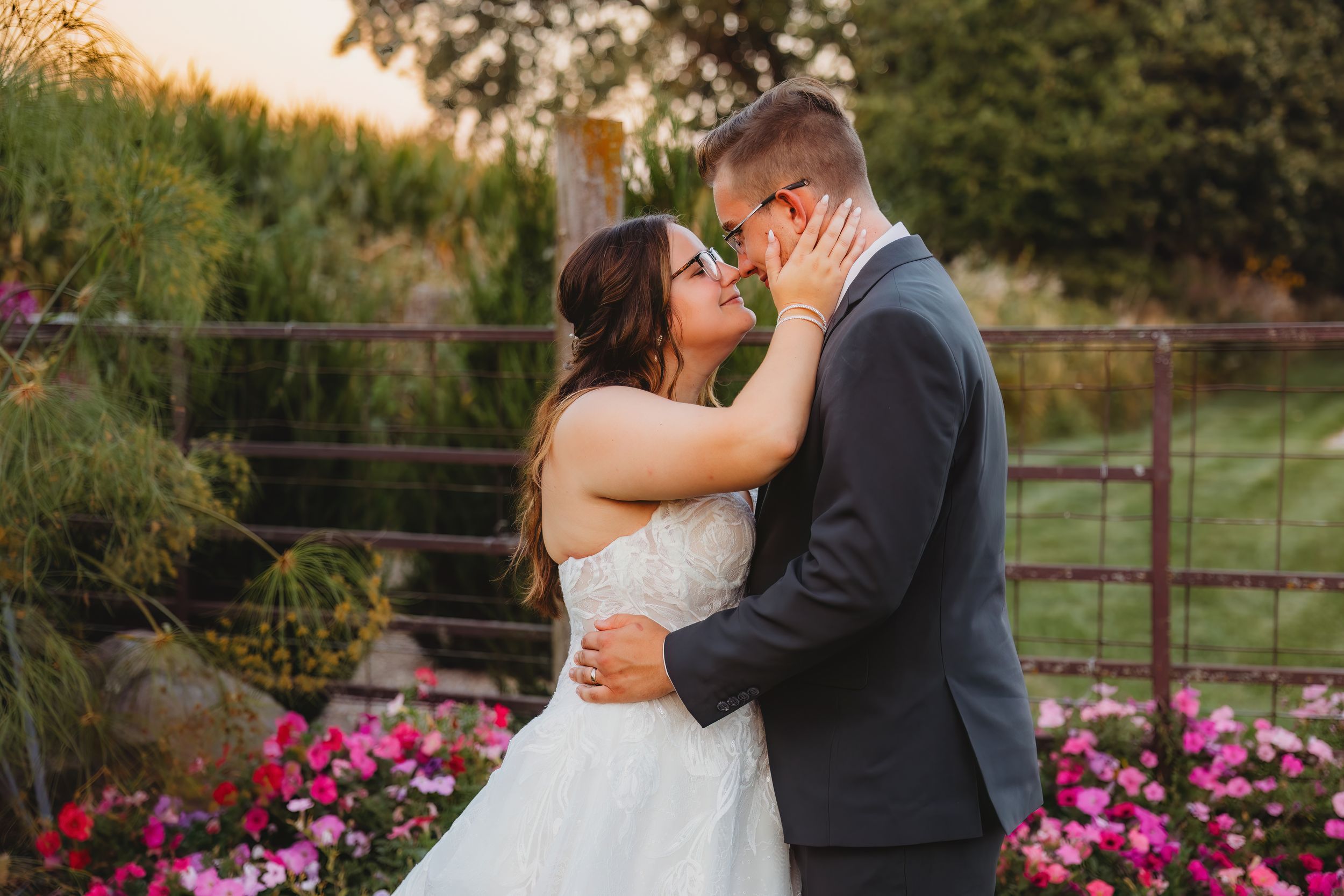 A couple shares a romantic moment in front of a garden with vibrant pink flowers during a sunset wedding ceremony.