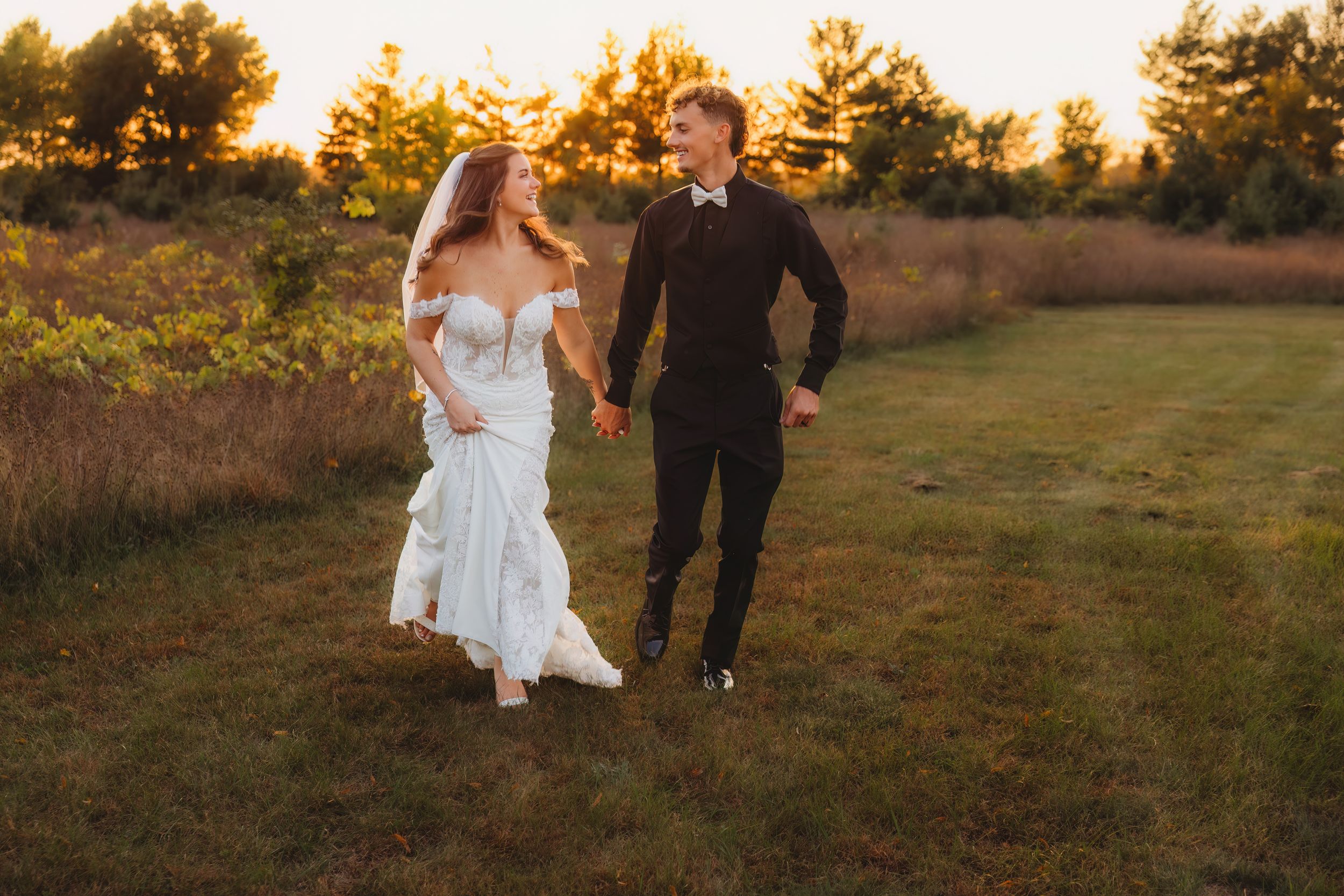 A couple walks through a golden field at sunset during their outdoor wedding photoshoot.