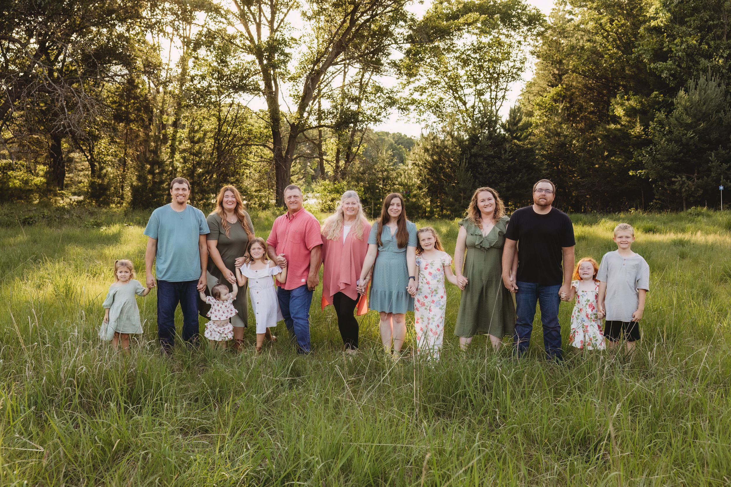 Large family group portrait outdoors in a lush green meadow with tall trees in golden hour sunlight.