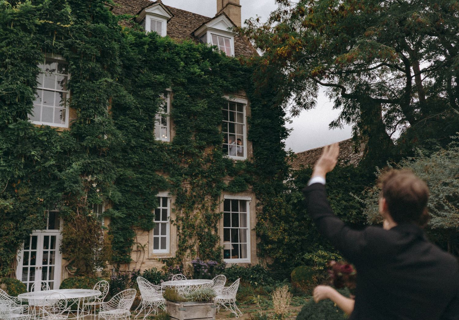 bride & groom waving at guest upstair through a window, cotswold wedding