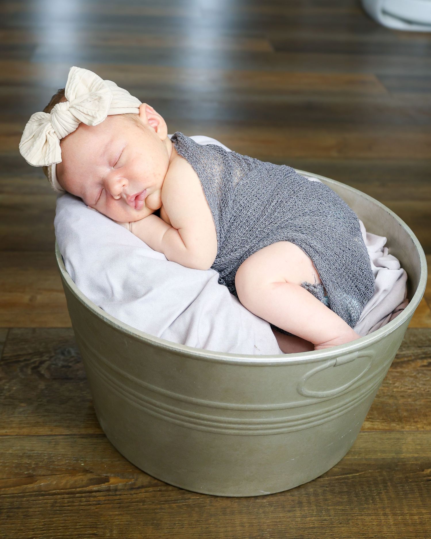 Sleeping newborn in gray outfit nestled in olive green bowl on wooden surface.