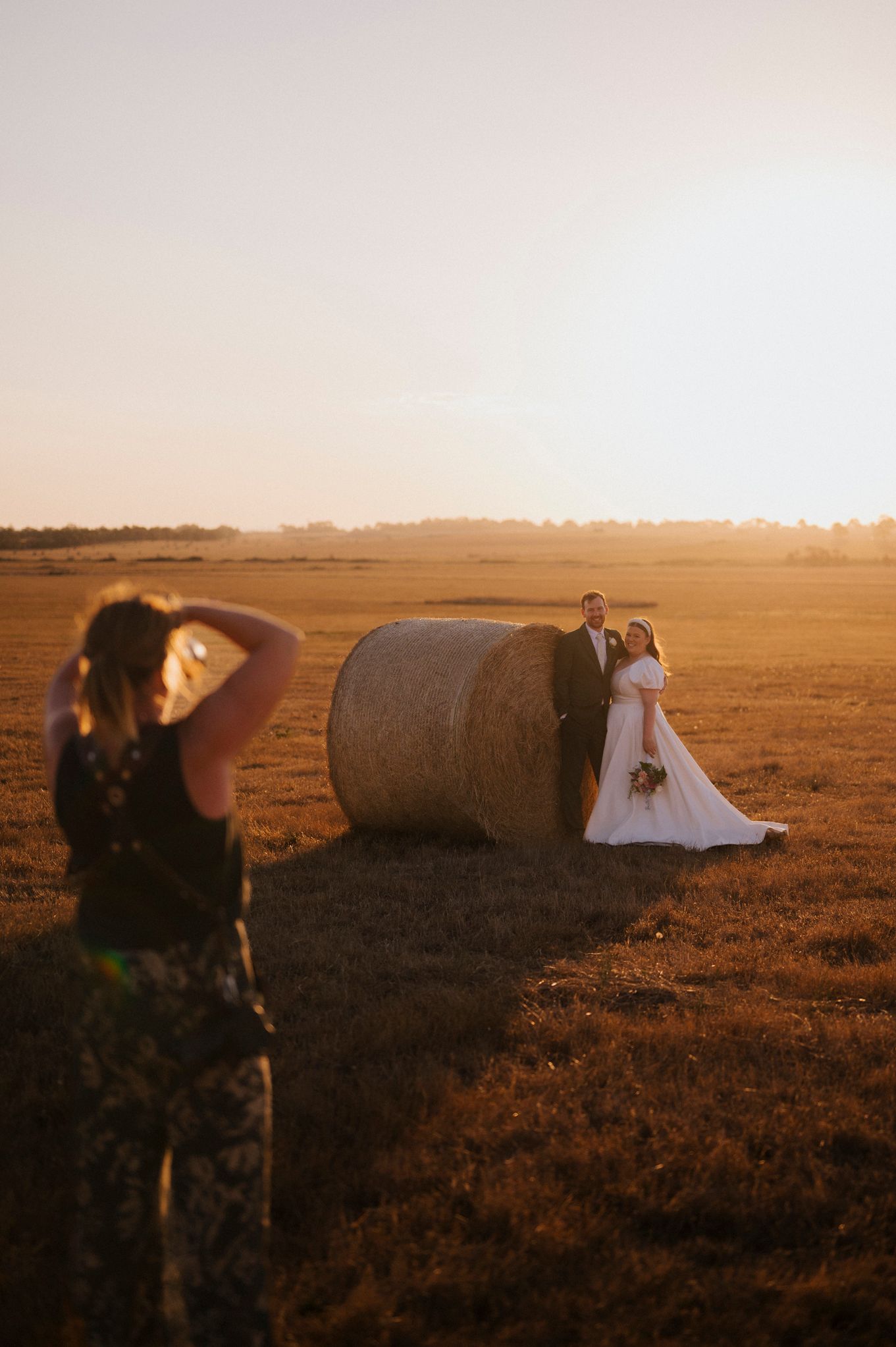 A wedding photographer captures couples posing near hay bales in a golden sunset-lit field during a rustic outdoor photoshoot.