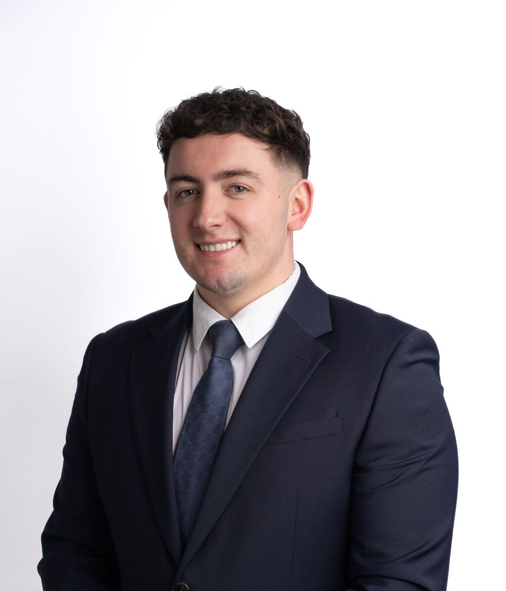 A friendly person in a navy suit and pink tie poses against a white background for a professional headshot.