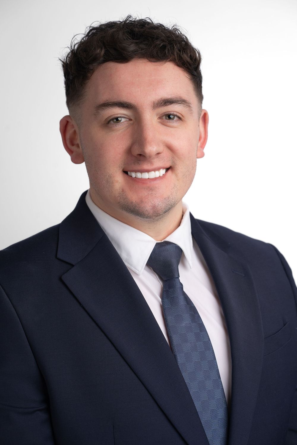 Professional headshot of a young professional in a navy suit and blue tie smiling against a light gray background.