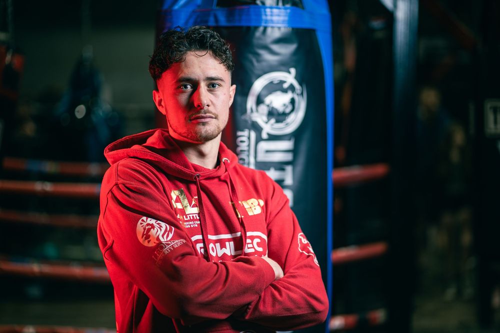 Athlete in red athletic jacket stands confidently in front of blue punching bag in dimly lit gym setting.