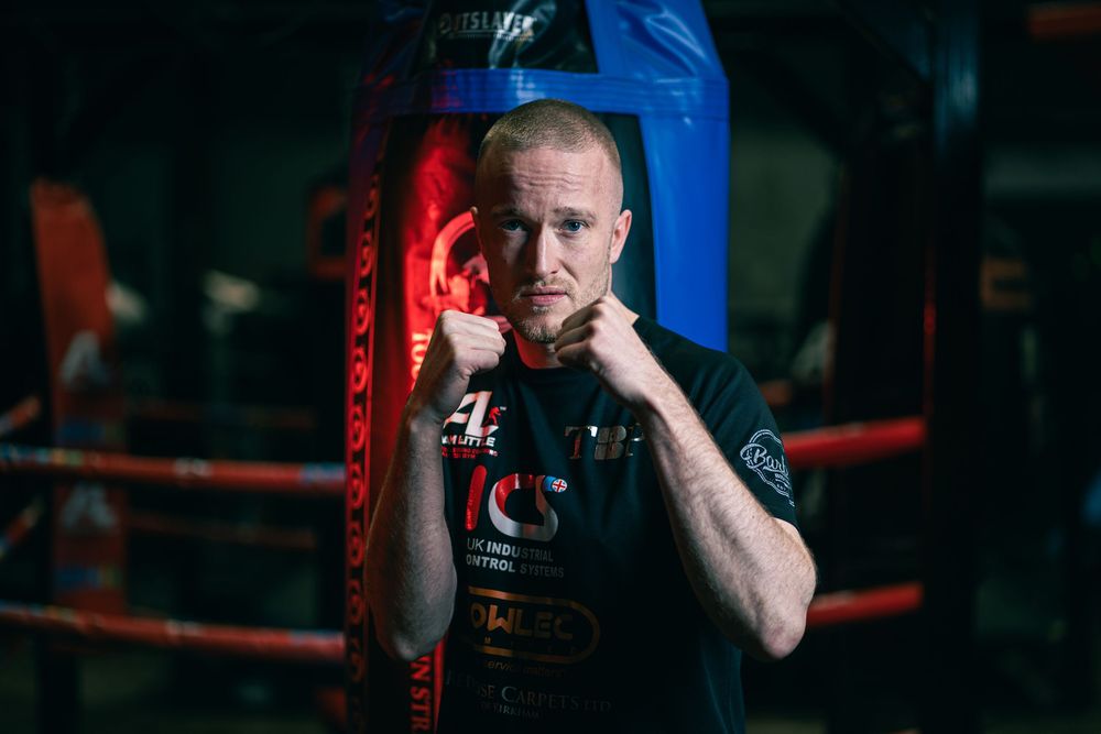 Boxer throws intense punches at heavy bag against blue and red lighting in dark gym setting.
