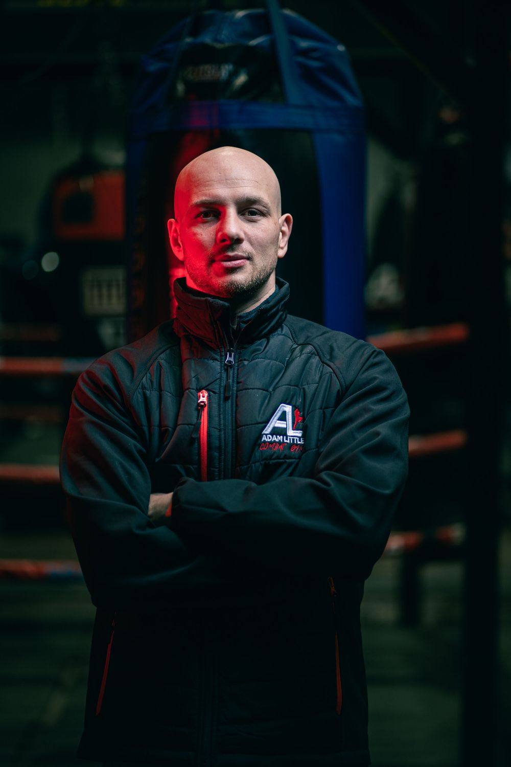 Person wearing black athletic jacket with red accents stands with arms crossed in dimly lit environment with red lighting.