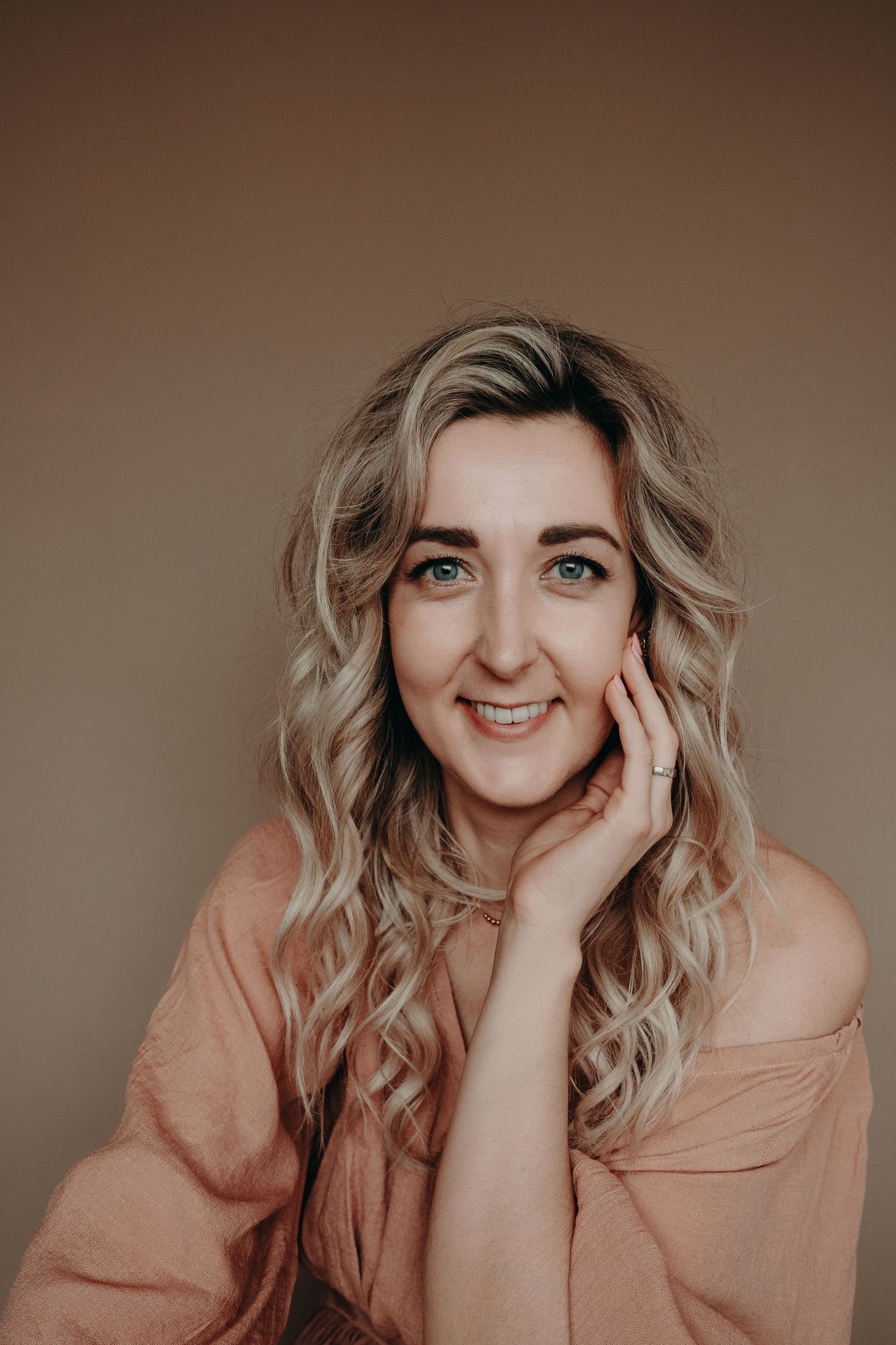 Portret tegen een bruine achtergrond van fotografe met golvend blond haar, gekleed in een perzikkleurige top en een zachte glimlach.