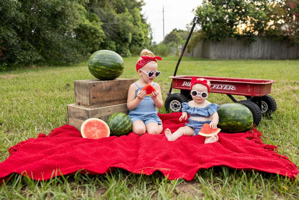 Summer picnic setup with watermelons and toys on a bright red blanket in green grass.