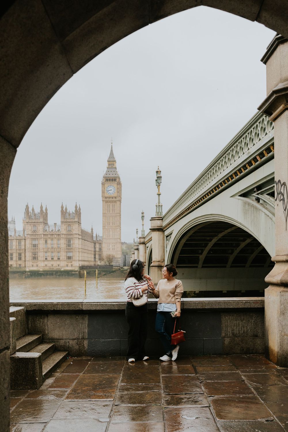 Two people sitting under Westminster Bridge with Big Ben and Houses of Parliament visible across the River Thames in London.