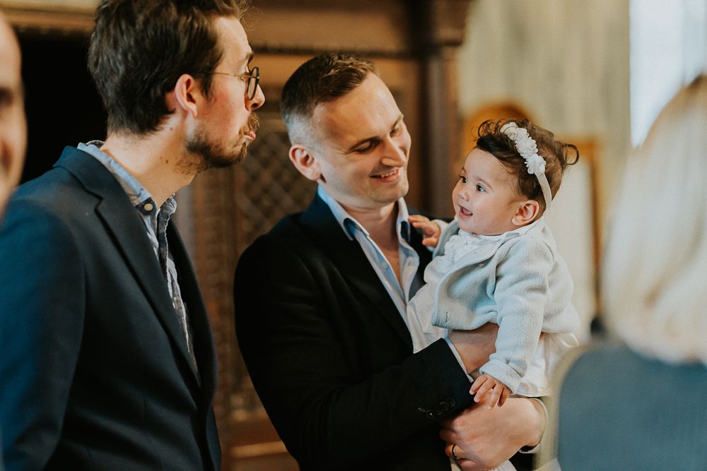 Two people in suits engaged in conversation while one holds a baby wearing a white bow headband and blue outfit.