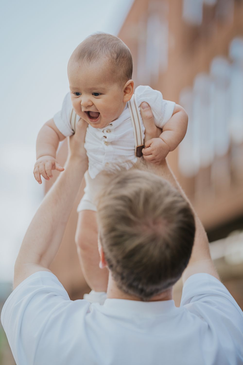 A happy baby in white overalls being lifted up in the air by someone in a white shirt on a sunny day.