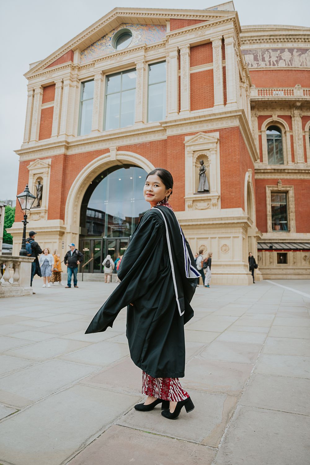 A graduate wearing academic regalia poses outside the Royal Albert Hall in London with its distinct pink architecture.