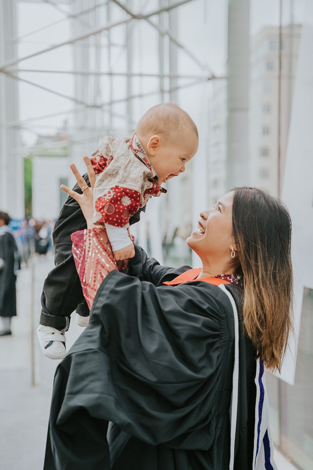 A graduate in cap and gown lifts up a baby wearing red and black on graduation day at university.