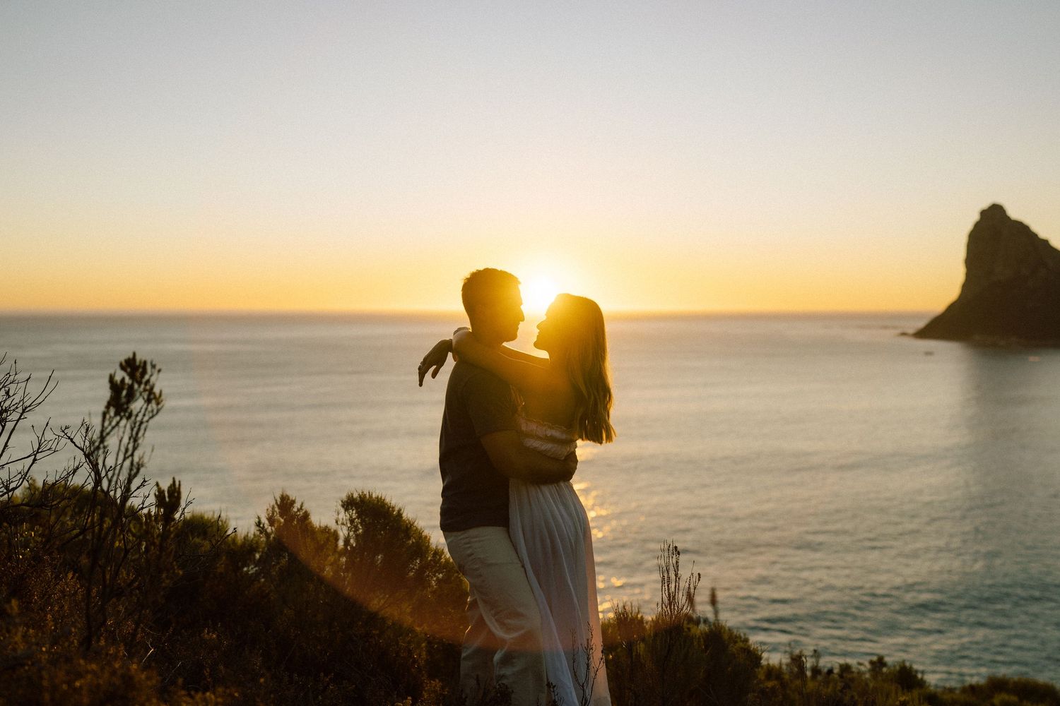 Silhouette of a couple standing on a hilltop against a dramatic sunset over a coastal city.