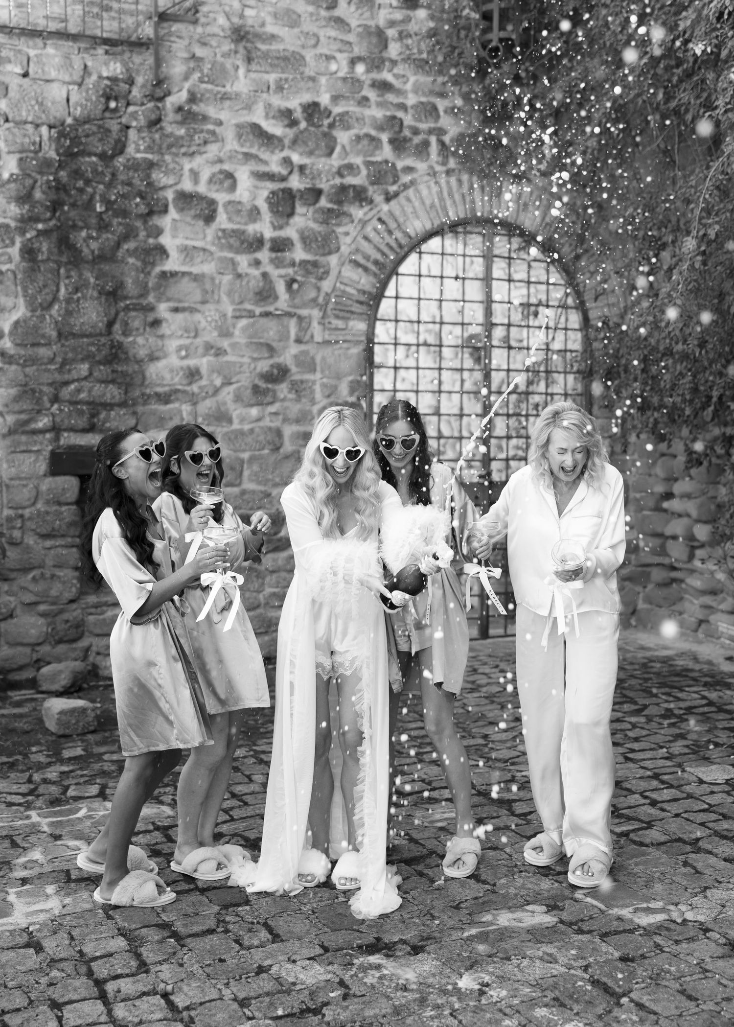 A group in white outfits stands laughing together on a cobblestone street in front of an old stone wall with an arched window.