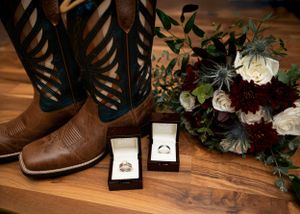 Wedding details with cowboy boots, wedding rings, and burgundy floral bouquet on wooden surface.